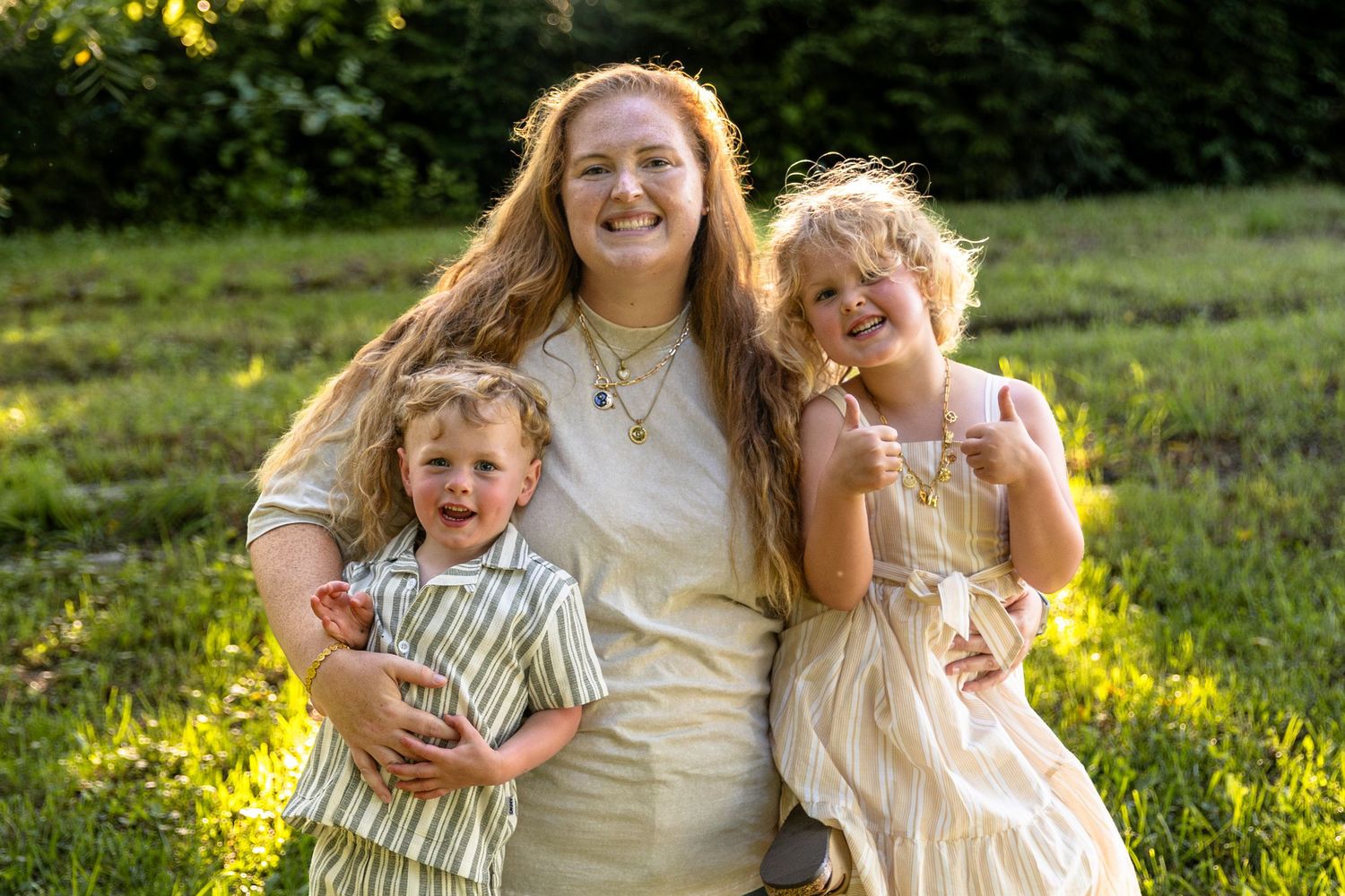 Smiling mother sitting outdoors with her two young children in golden sunlight.