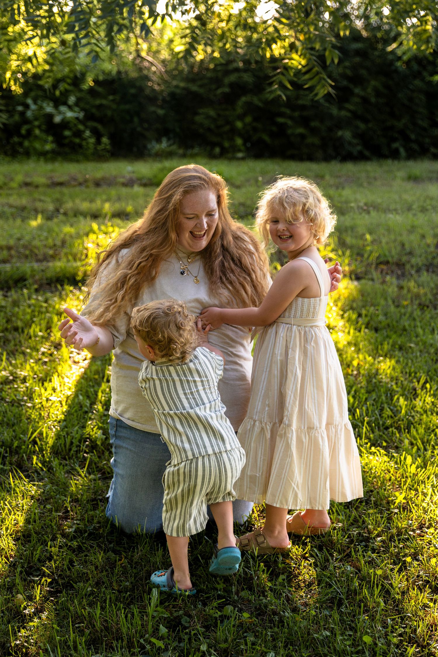 A laughing mother with long red hair plays with her two young children on a sunny green lawn.
