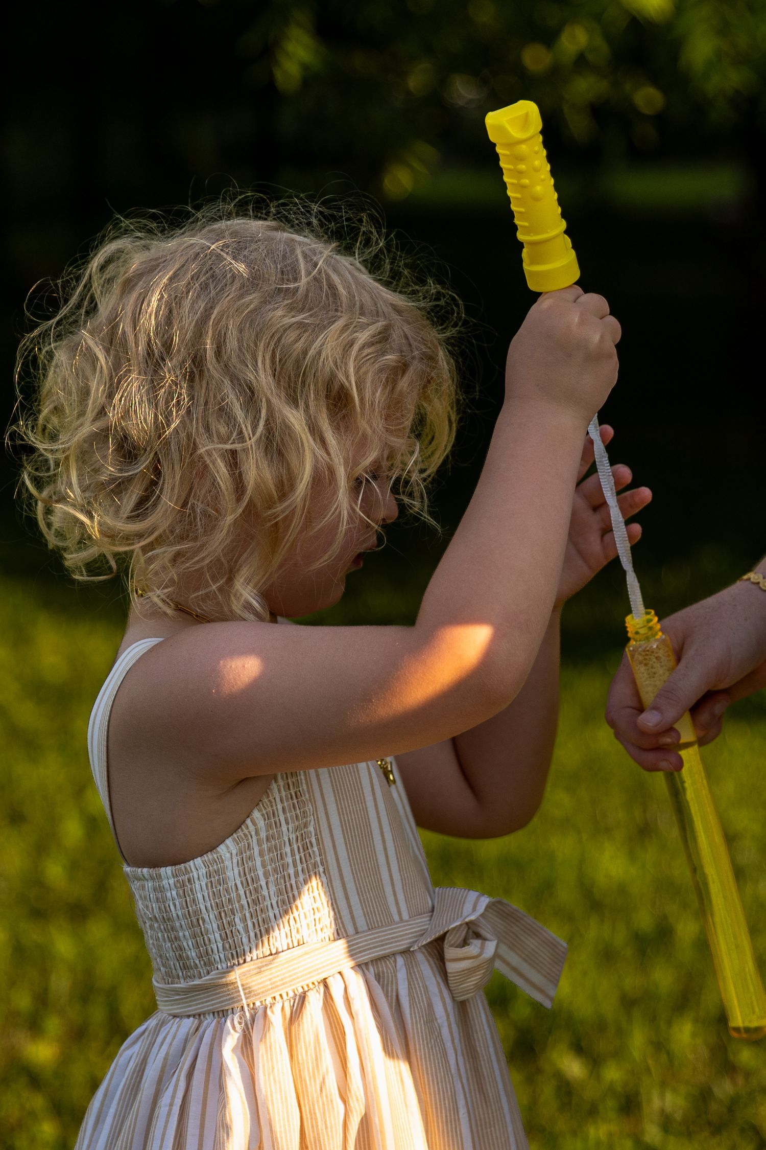 A young blonde curly-haired girl in a striped dress plays with a yellow bubble wand outdoors.