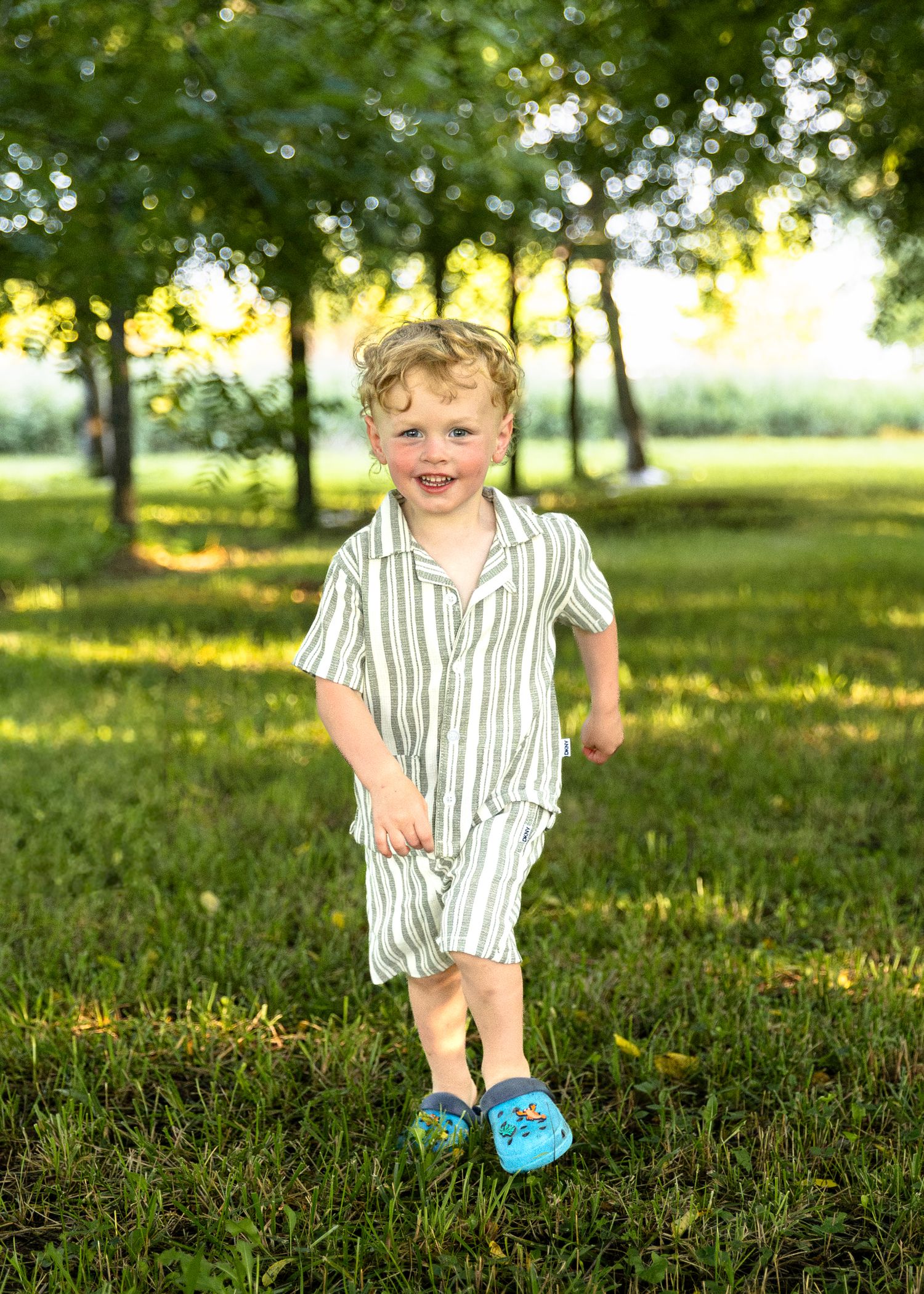Happy curly-haired toddler boy in striped outfit and blue crocs running through a sunny green park.