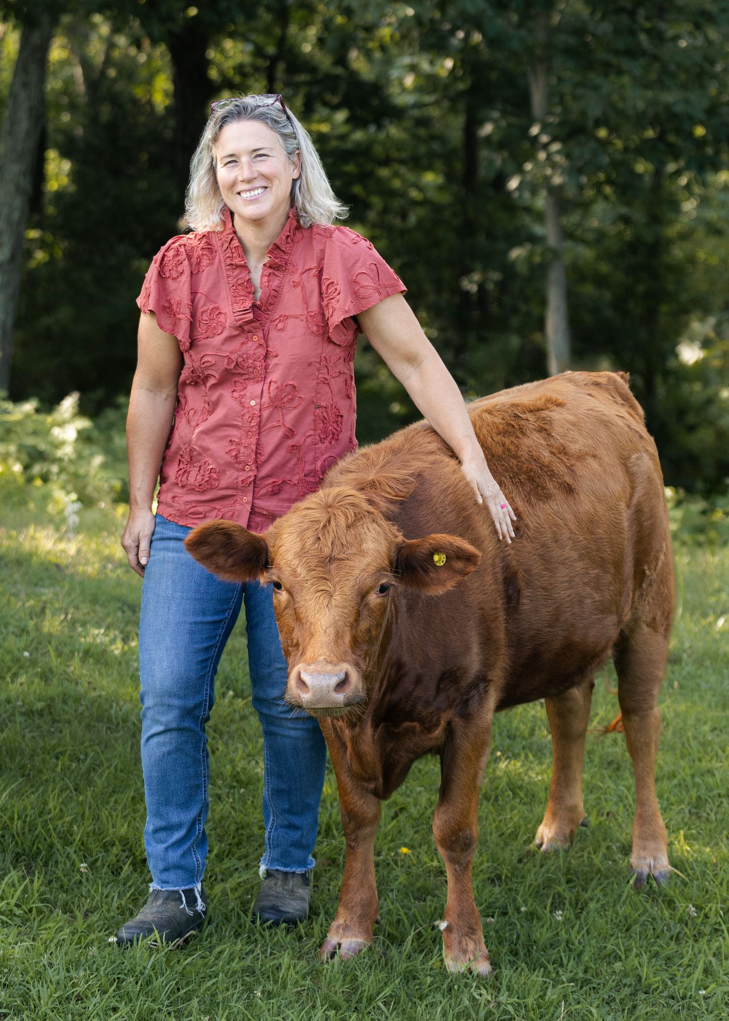 Woman in red blouse smiling beside a brown calf on green grass farmland.