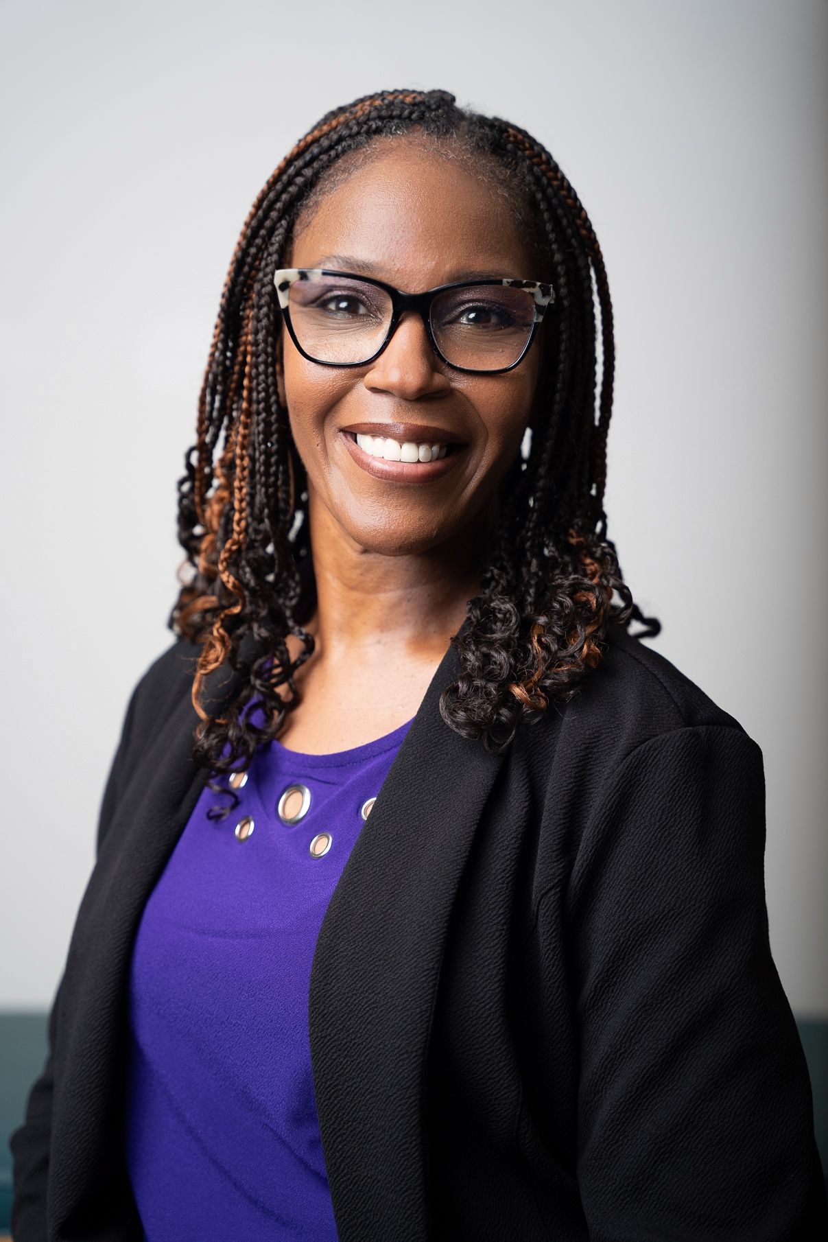 Professional headshots of person in black blazer and purple top wearing glasses.