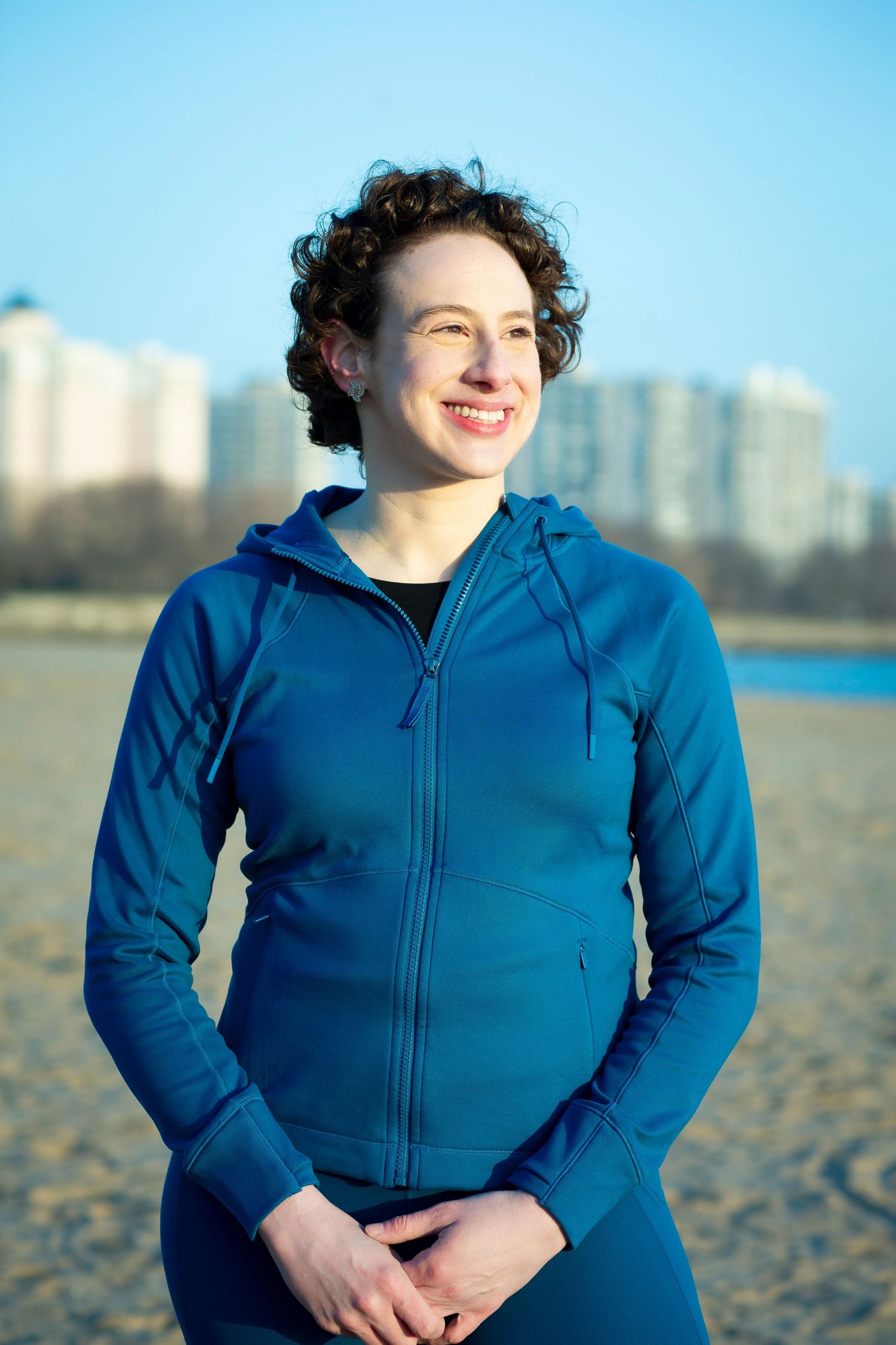 Person wearing a blue zip-up hoodie smiles while standing on a beach with city buildings in the background.