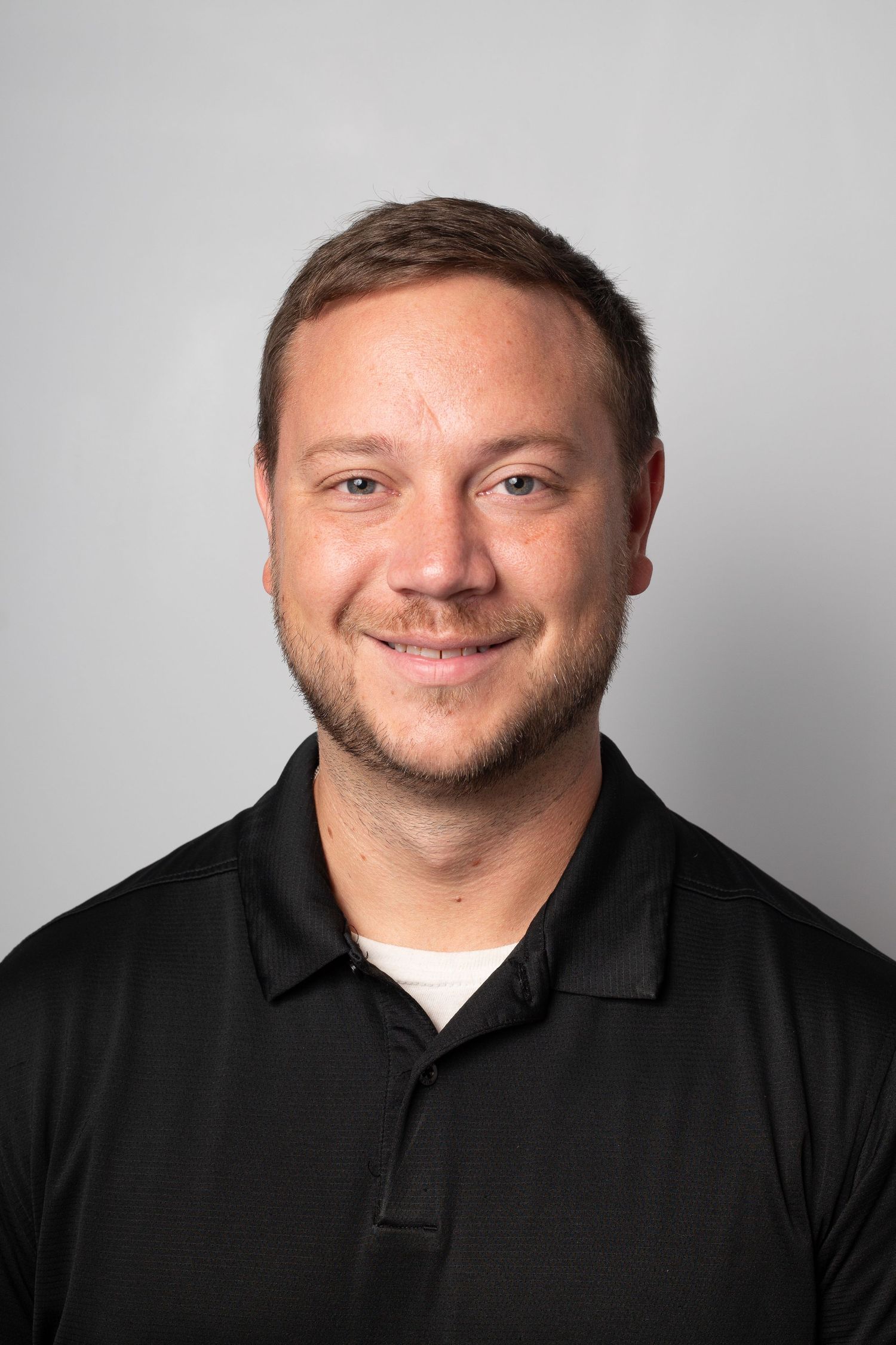 Professional headshot of a person wearing a black polo shirt smiling against a light gray background.