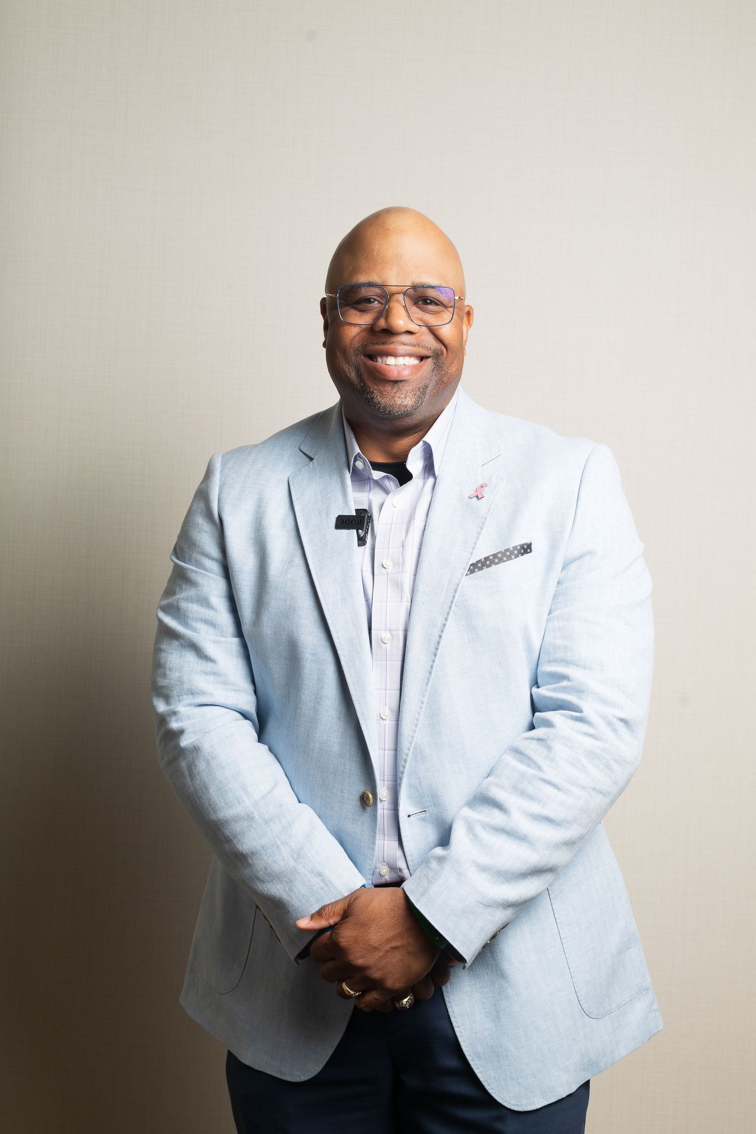A person wearing a light blue blazer and glasses smiles while standing against a neutral background.