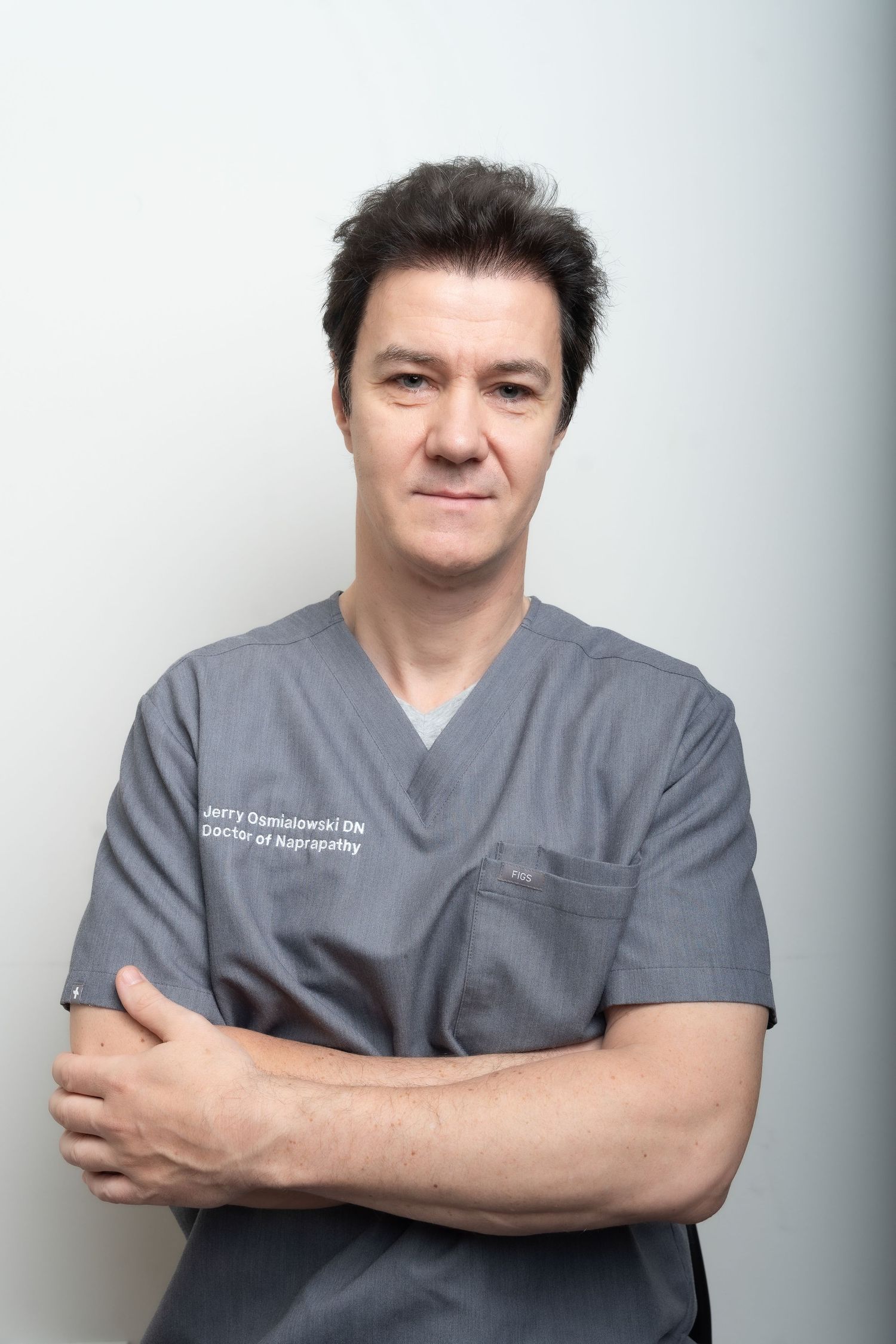 Healthcare professional in gray scrubs poses with arms crossed against light background.