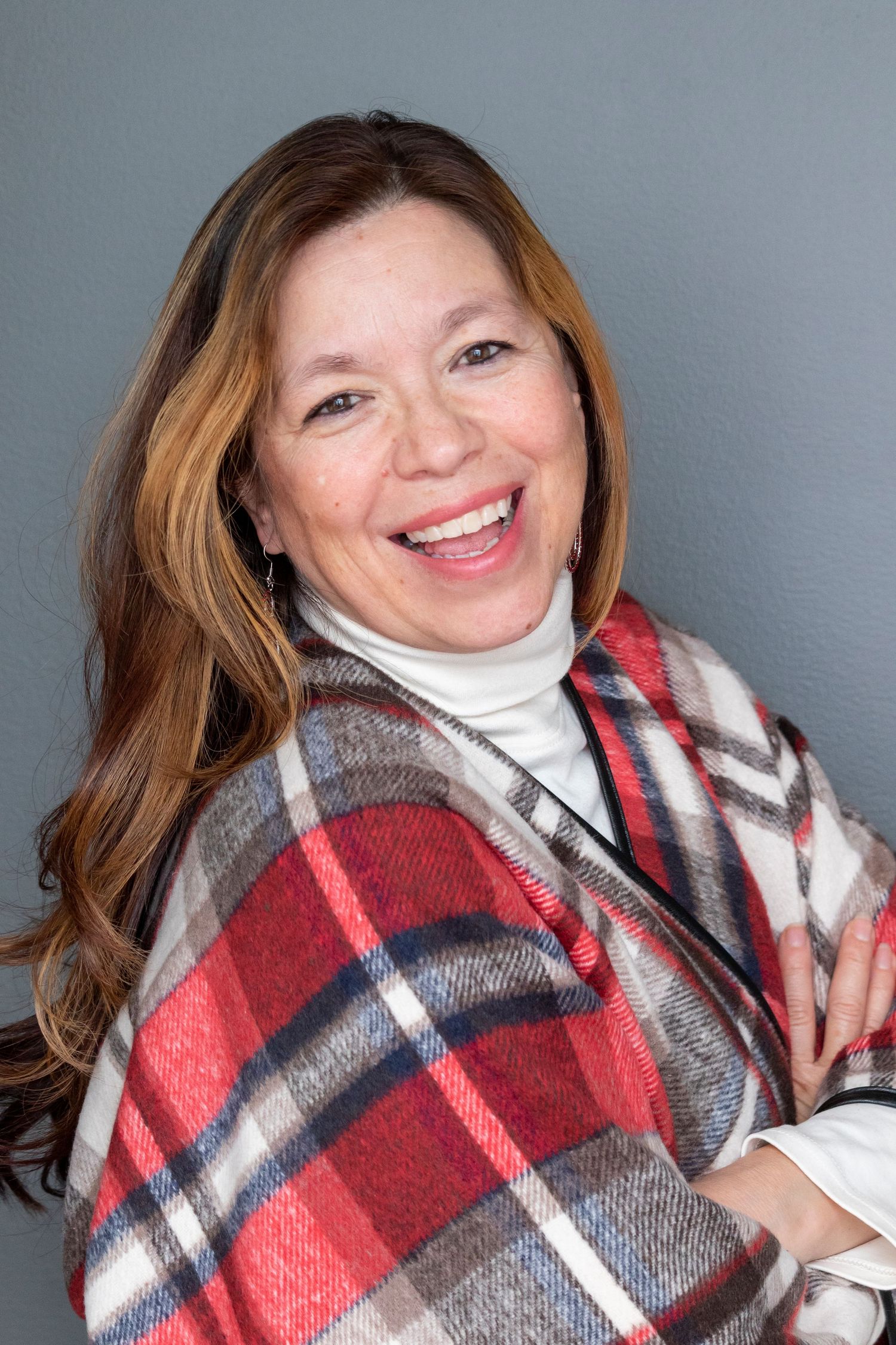 Person wrapped in a cozy red, white and gray plaid blanket smiling at camera against gray background.