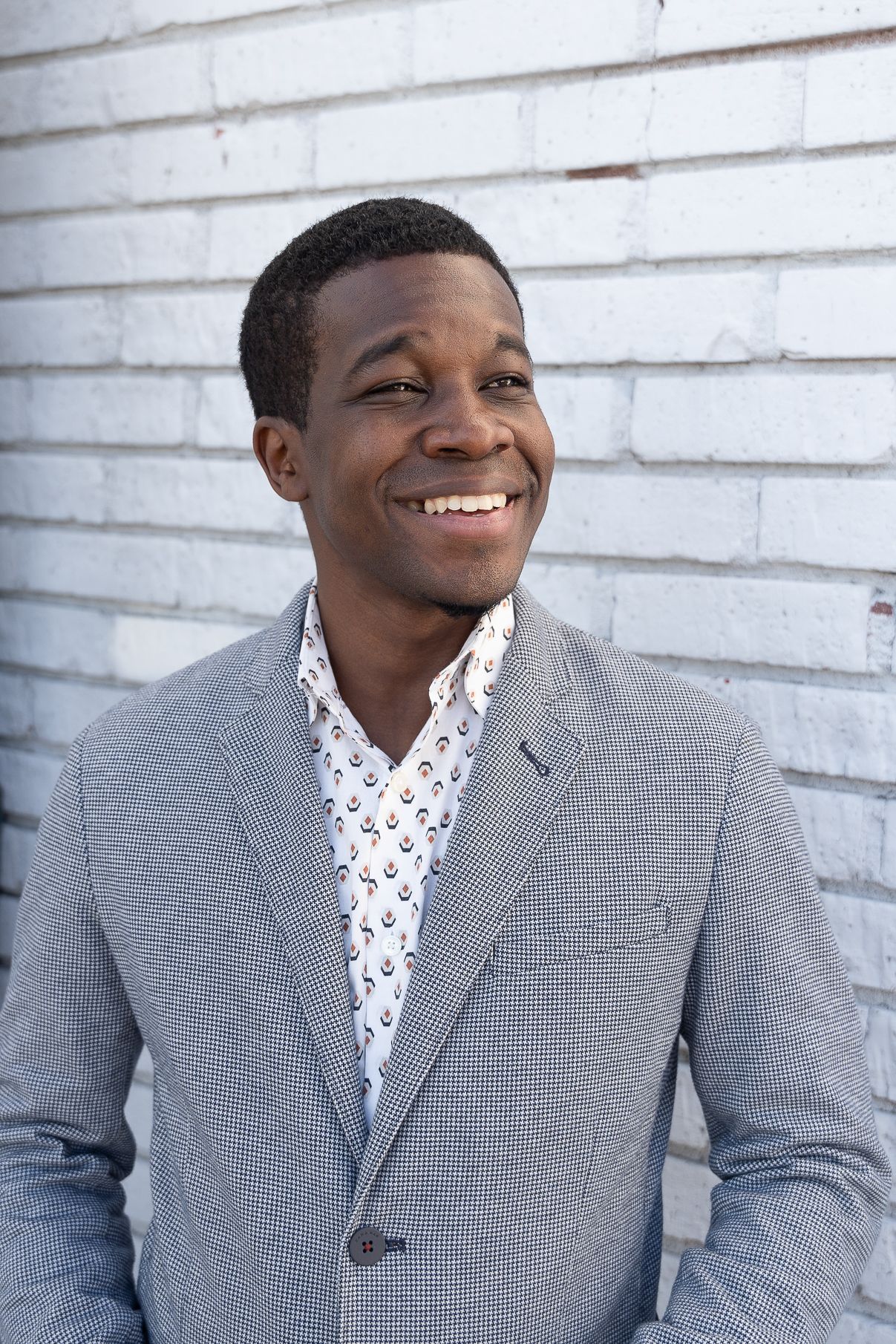 A person in a gray suit jacket and patterned white shirt poses against a white brick wall in a progression of expressions.