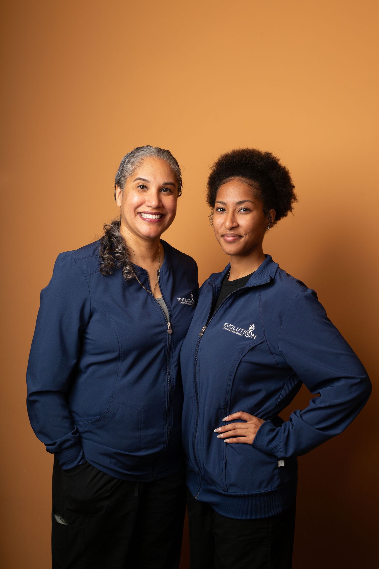 Two people wearing matching navy blue uniforms pose together against an orange backdrop in a professional photoshoot.