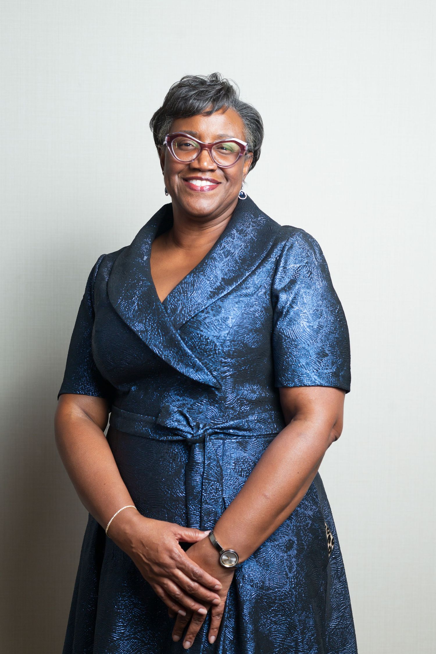 A person wearing a blue brocade wrap dress and glasses poses confidently against a white background.