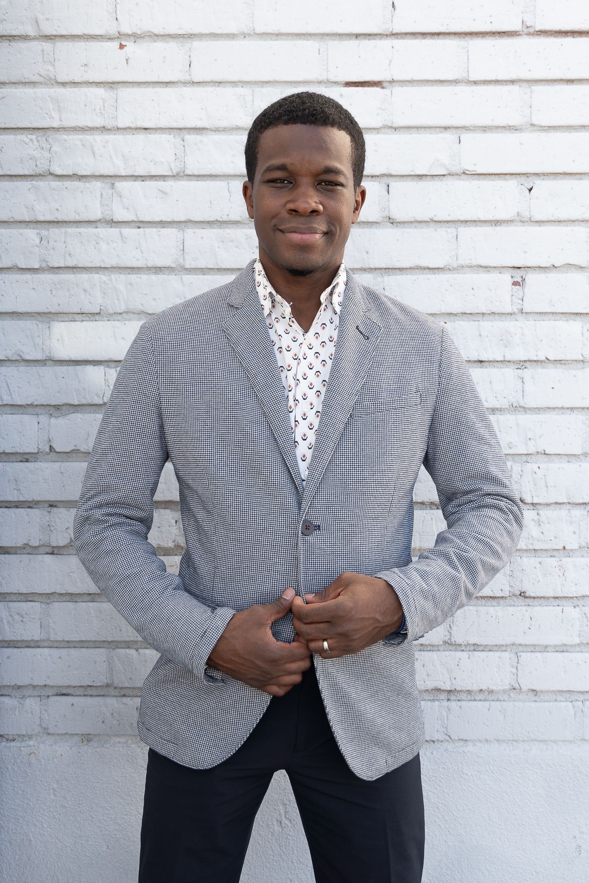 Professional portrait against white brick wall showing gray blazer and polka dot shirt styling.