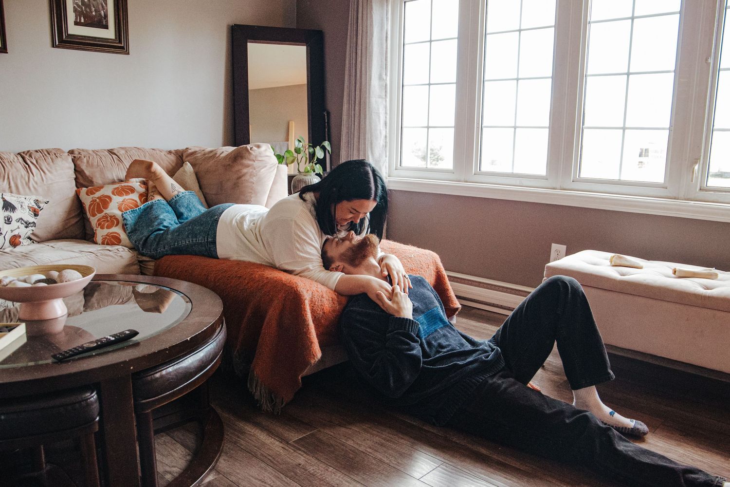 She leans in to kiss her fiancee on their cozy couch with natural light spilling into the room
