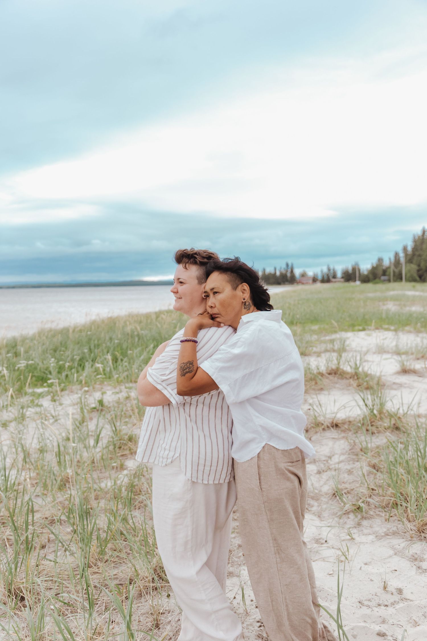 A couple stands on the shores, leaning on one another watching the waves roll in