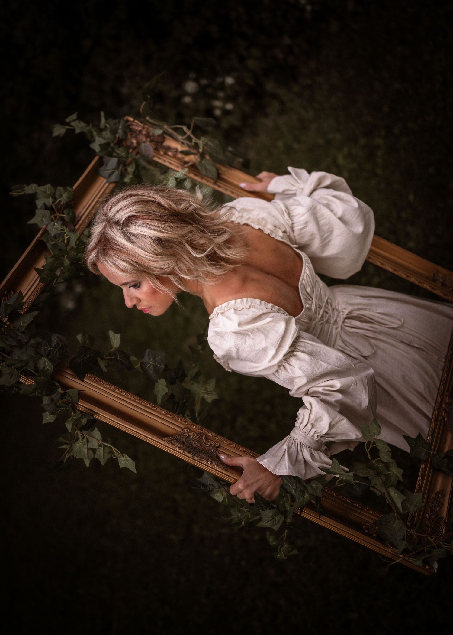 Blonde woman in white off-shoulder dress leaning through ornate gold frame adorned with ivy leaves outdoors.