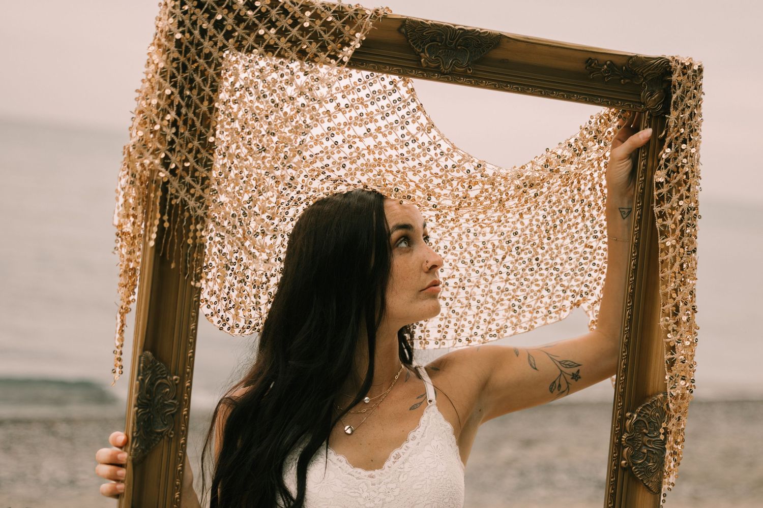 Woman holding wooden frame with gold chain mesh at beach, wearing white dress with tattoo visible.