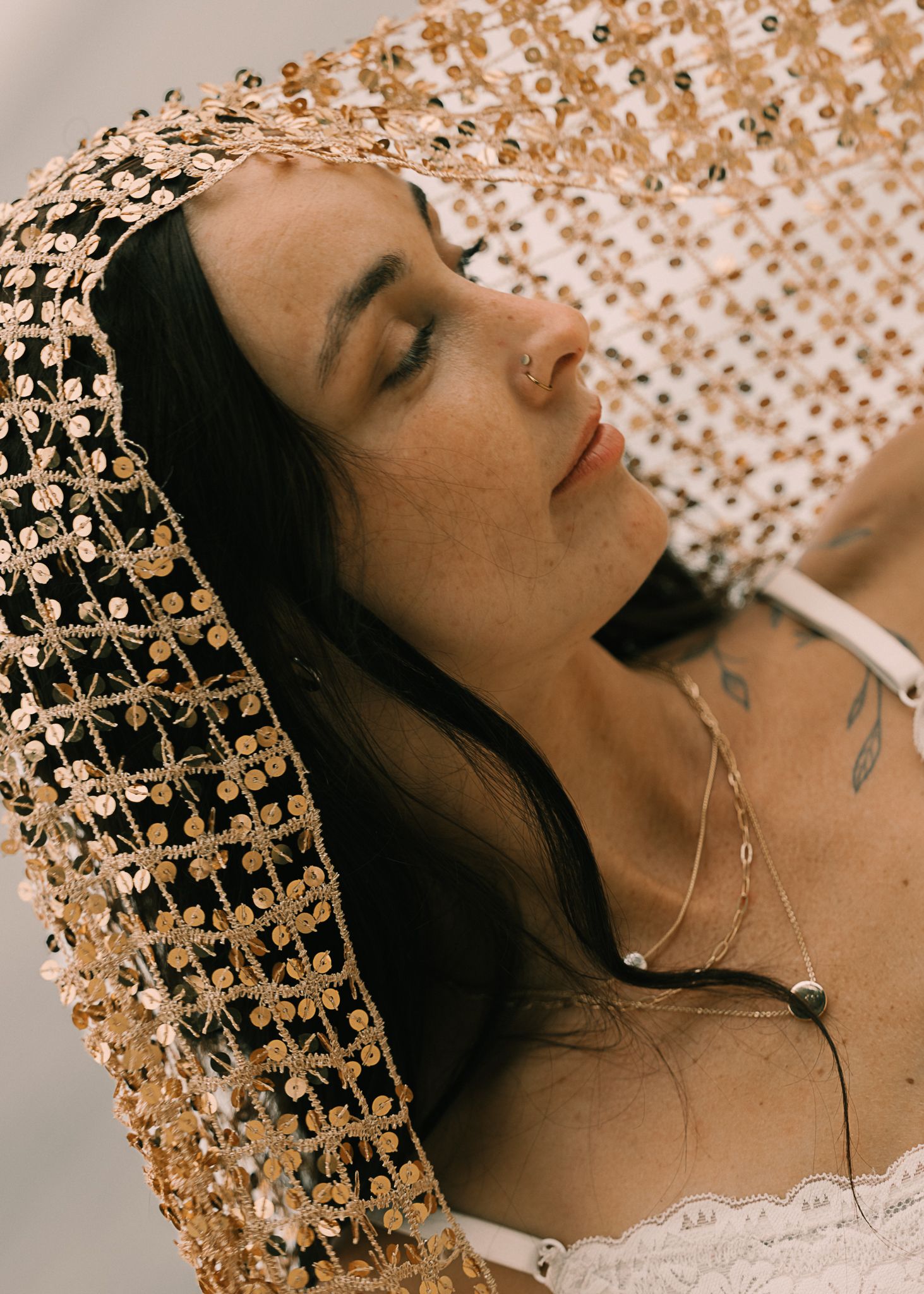 Woman wearing a gold sequined veil with floral embroidery, eyes closed, wearing a delicate necklace and lace top.
