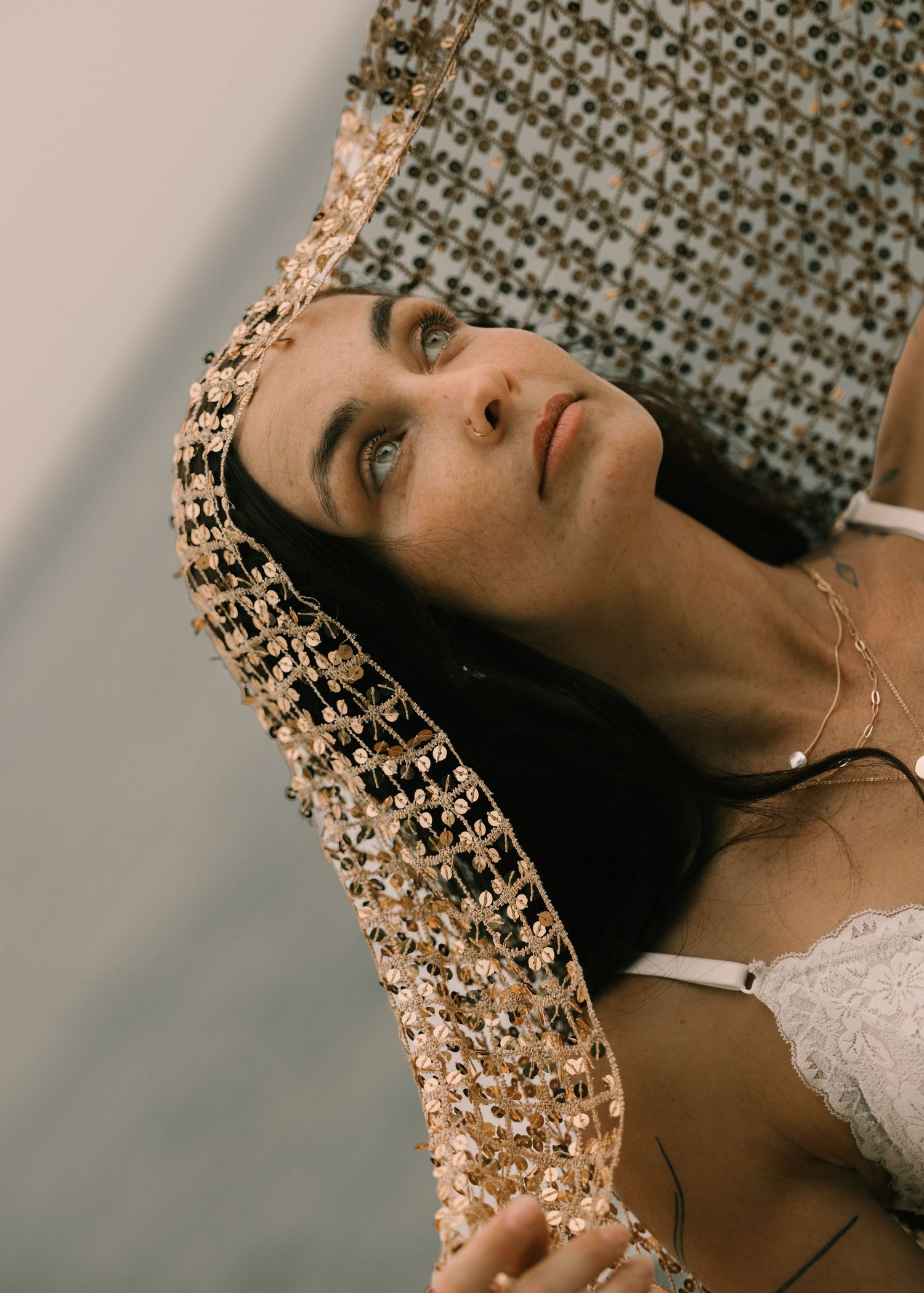 Woman lying down draped in gold sequined fabric, wearing white lace top and delicate necklace, gazing upward.