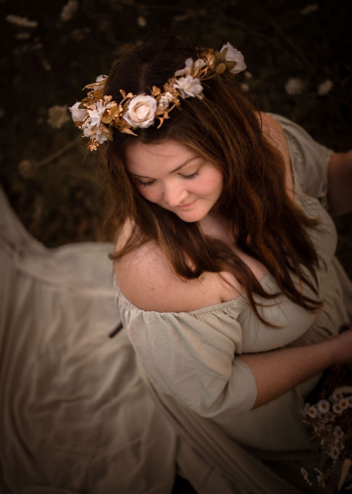 Young woman in off-shoulder dress wearing gold and white floral crown, smiling softly in an outdoor portrait.