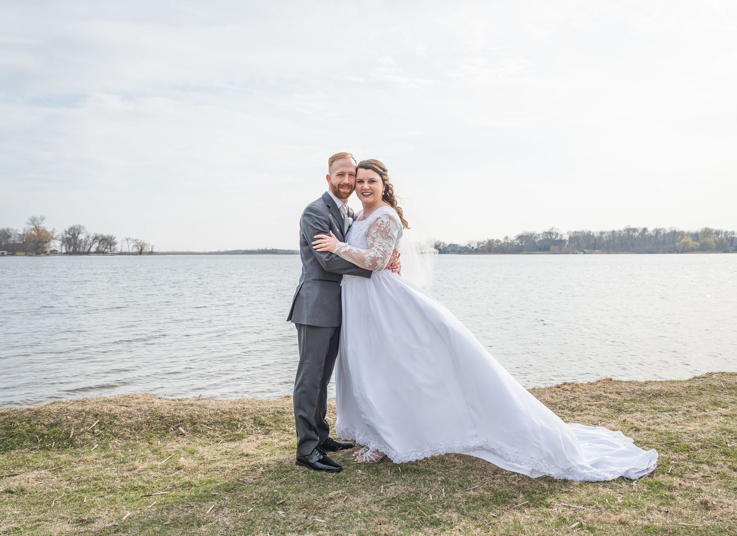 Smiling red-bearded groom in gray suit with bride's lace-sleeved hand gently touching his face on wedding day.