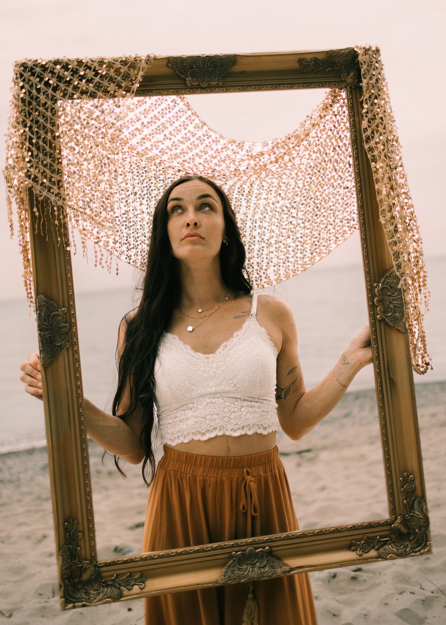 Woman in white crop top and gold skirt holds ornate picture frame on beach.