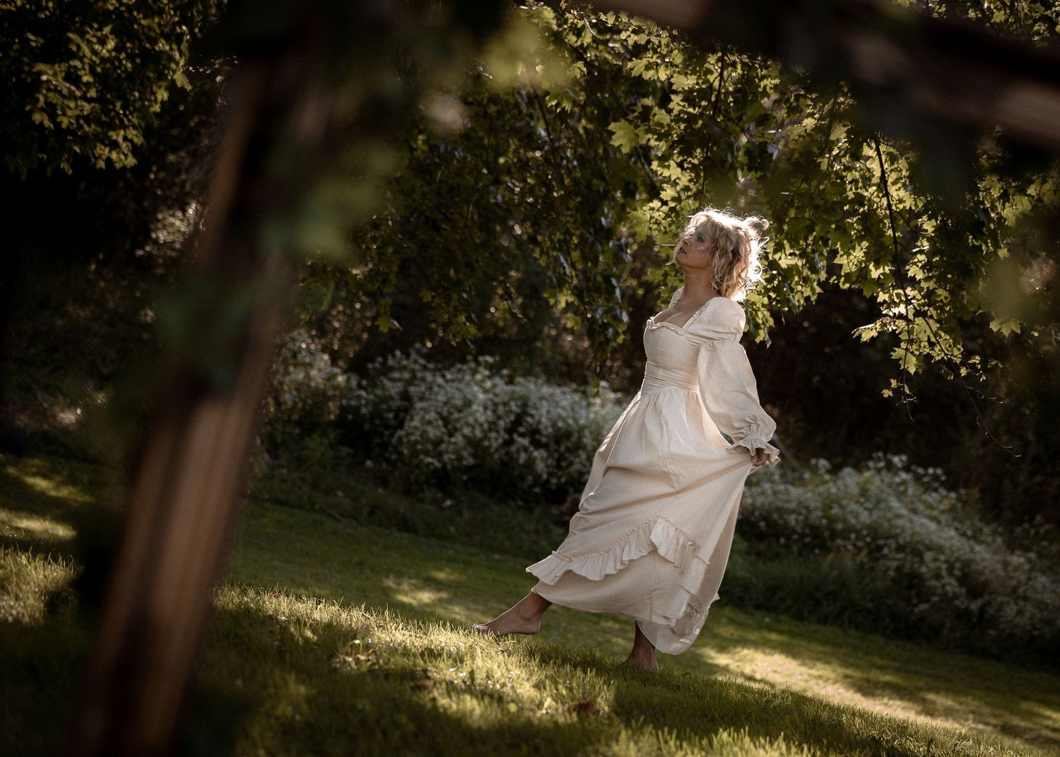 Woman in flowing white dress dancing gracefully in sunlit garden surrounded by lush green trees.