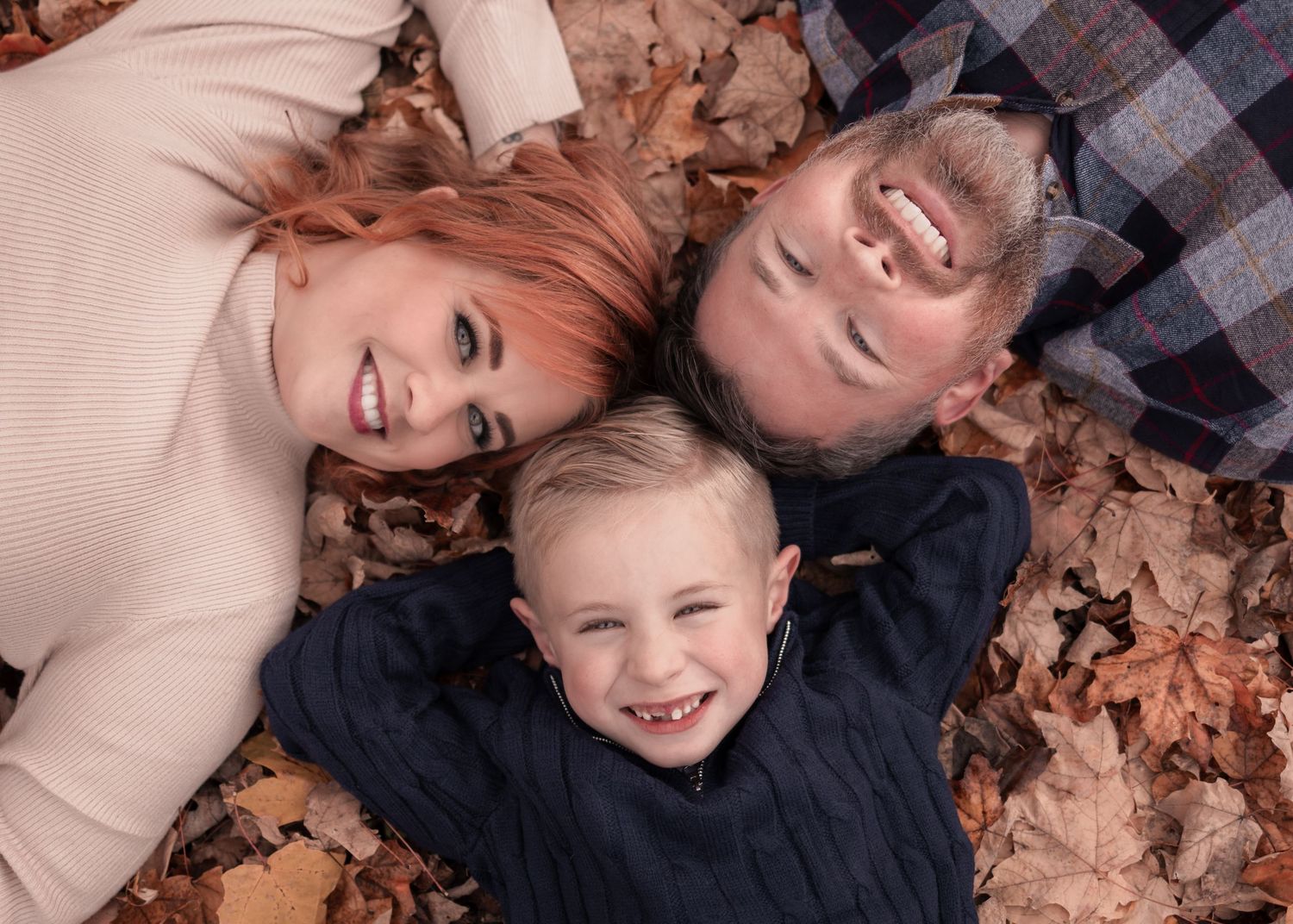 Happy family of three smiles lying together surrounded by golden fall leaves outdoors.