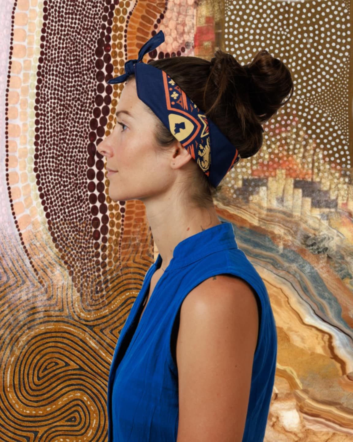 Woman in blue sleeveless dress viewed in profile standing before a colorful Aboriginal dot painting artwork.