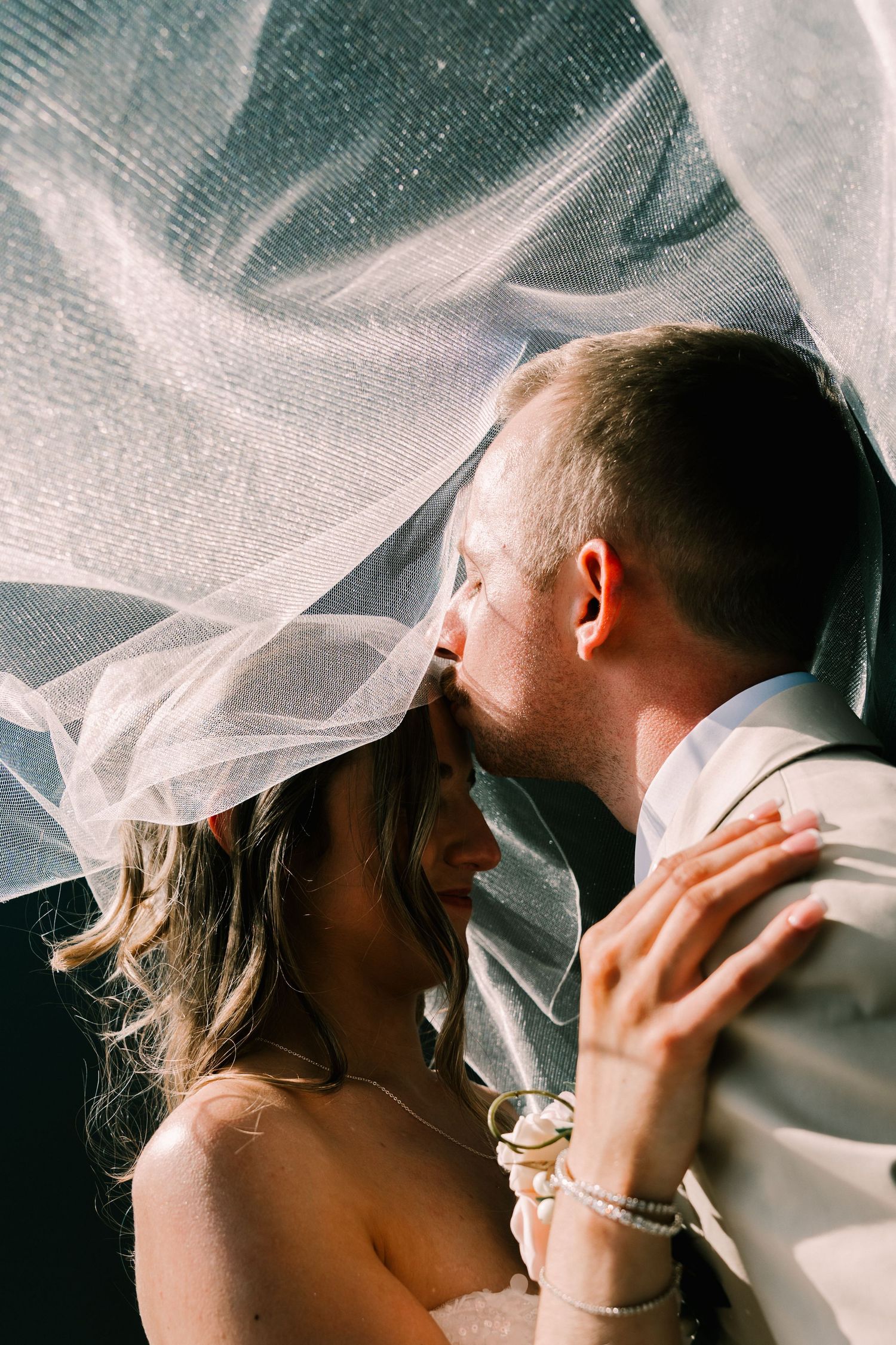 Bride and groom sharing a romantic kiss under a flowing white wedding veil, with the bride holding a champagne glass.