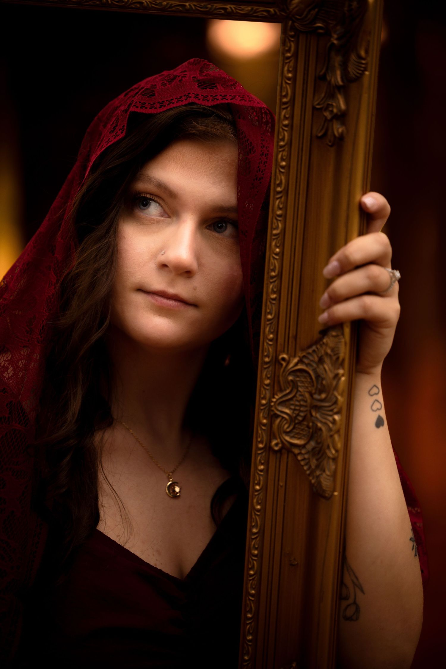 Young woman with red lace hood holding ornate gold picture frame, tattoos visible, wearing pendant necklace.