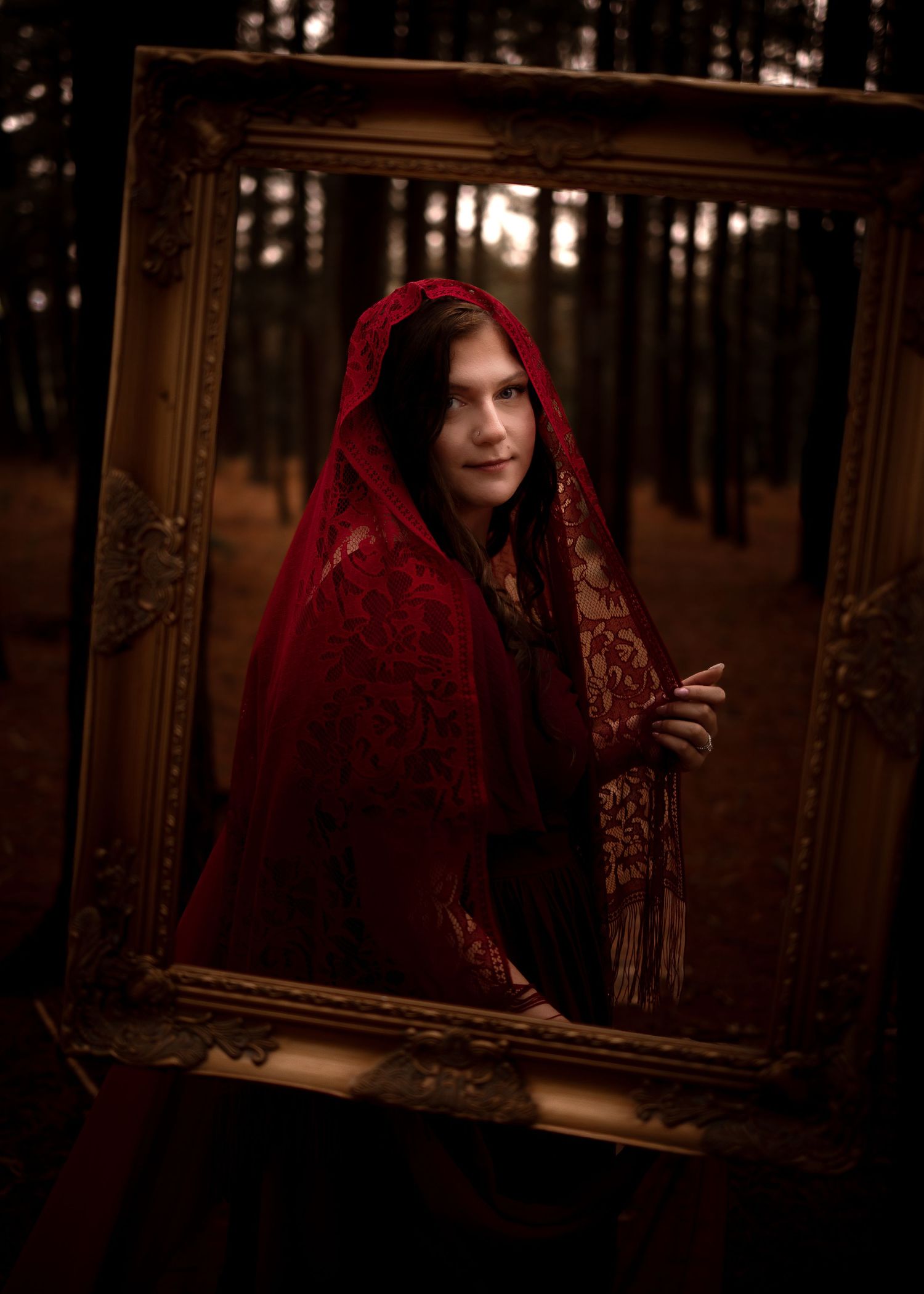 Person in red lace hood holding gold frame in dark forest, dramatic fairytale portrait photography.
