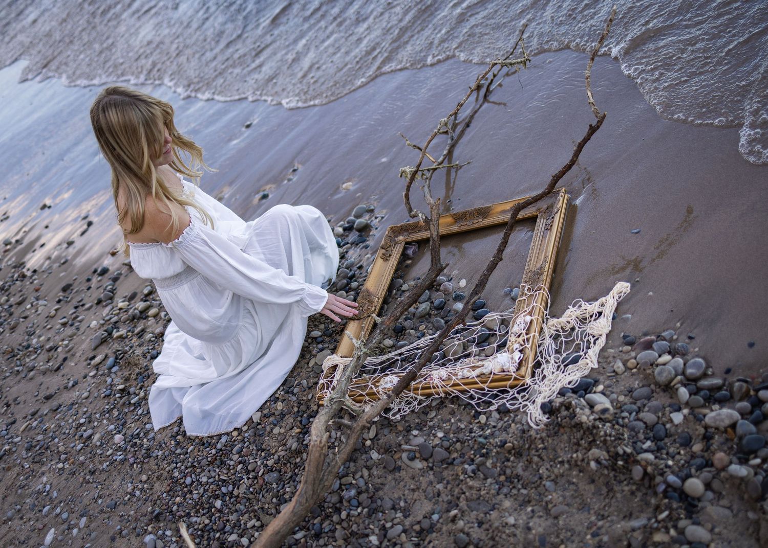 Woman in white dress sitting on rocky beach beside driftwood and a broken frame with waves in background.