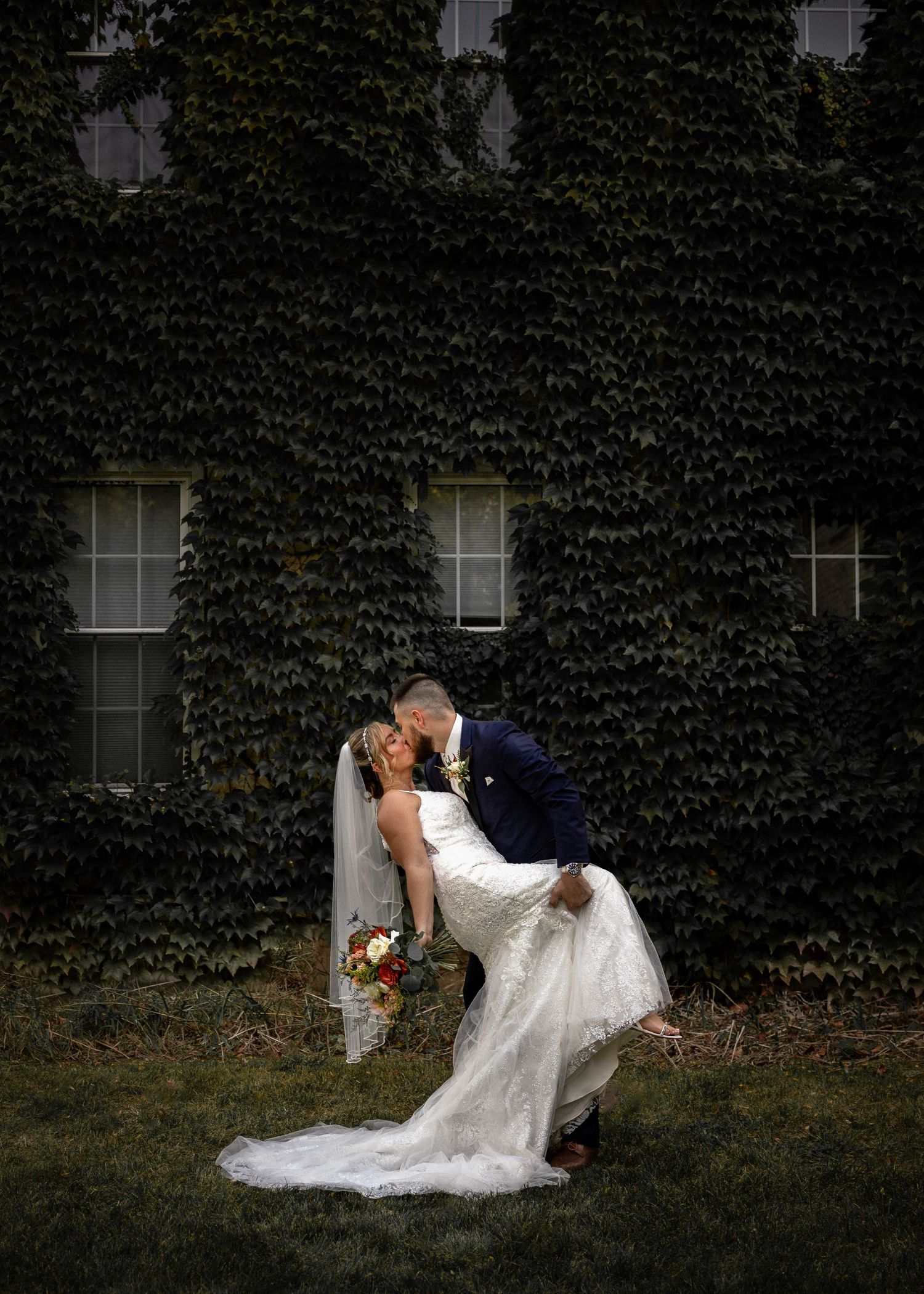 Bride and groom sharing romantic dip kiss in front of ivy-covered building wall.