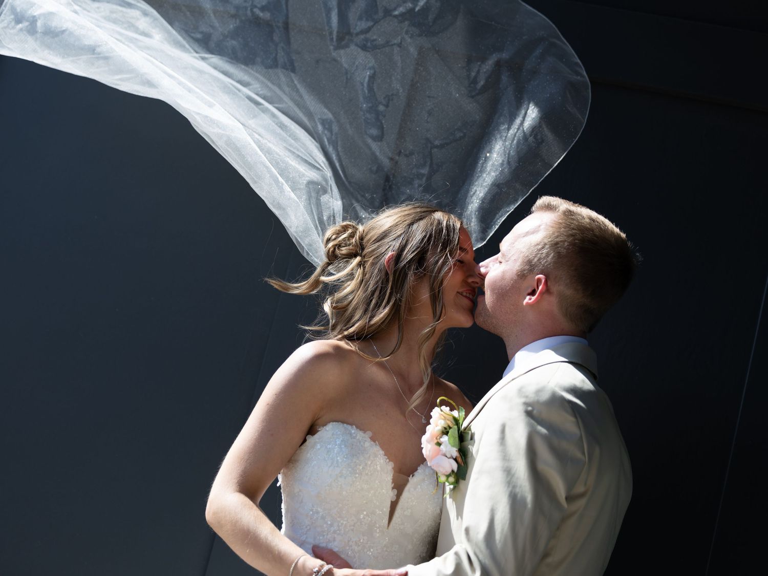 Romantic outdoor wedding kiss with bride's veil dramatically blowing in the wind against dark background.