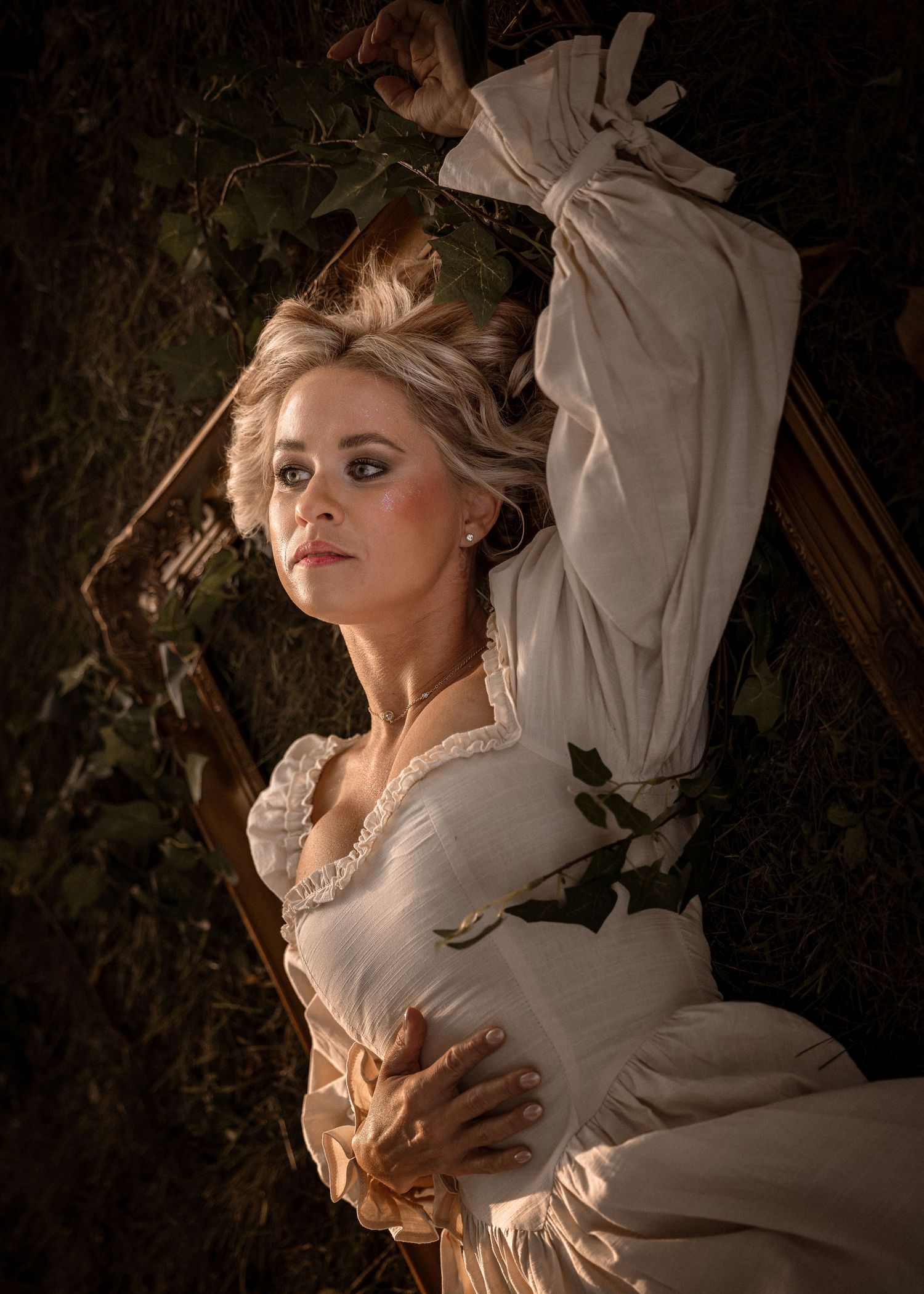 Woman in white period dress poses dramatically against ivy-covered backdrop in moody, theatrical portrait photography.