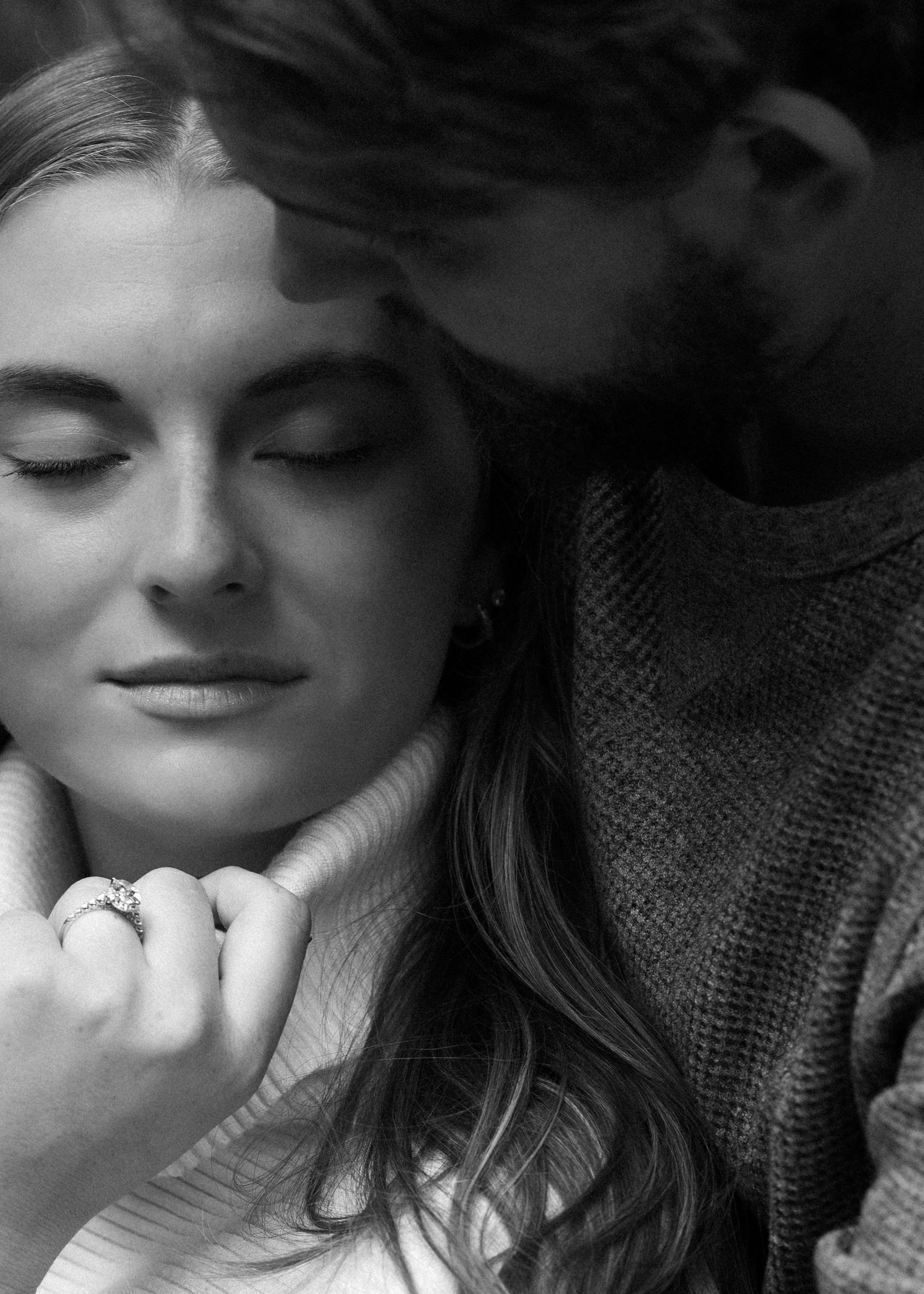 Woman with eyes closed wearing diamond ring as man embraces her in black and white.