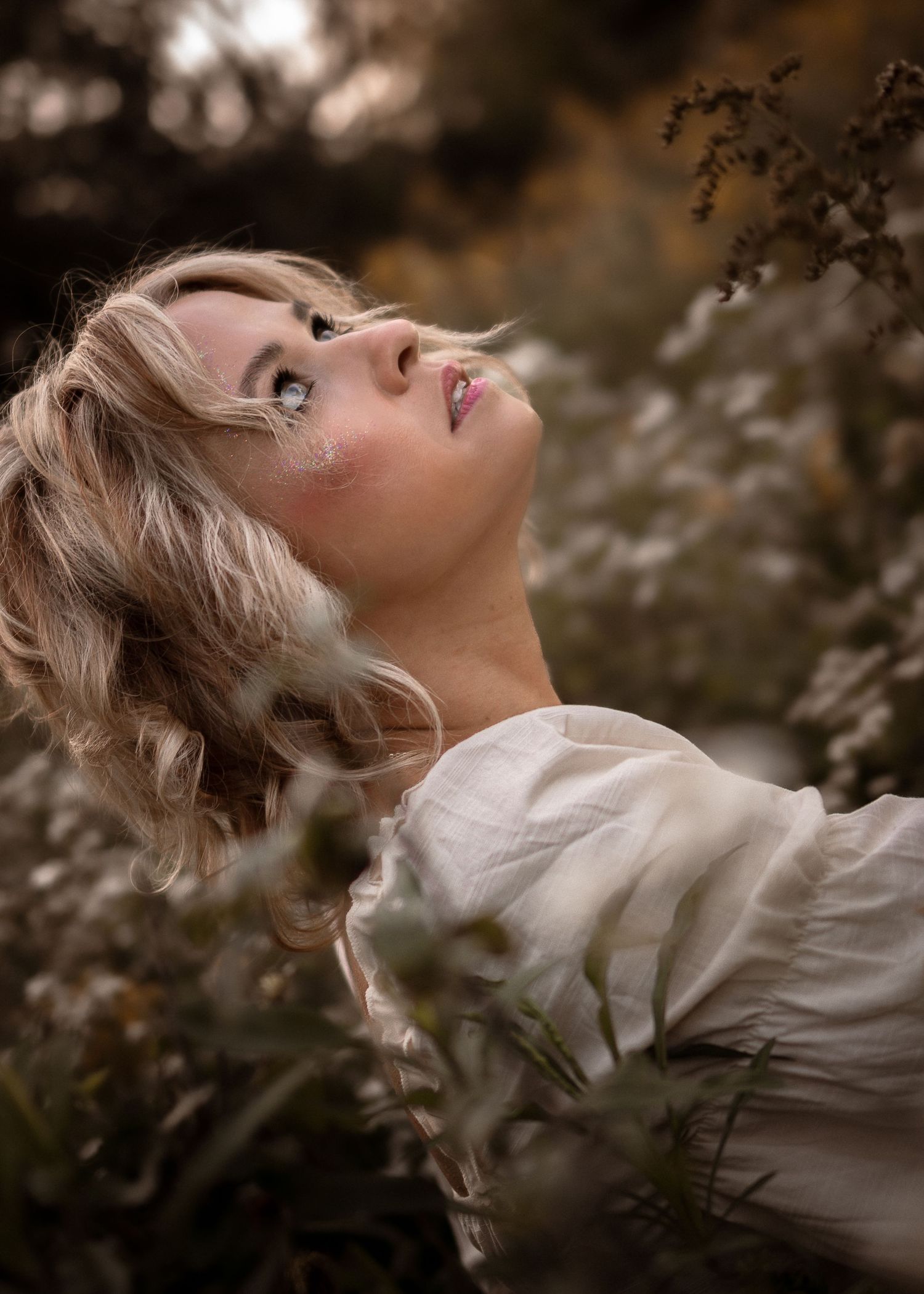Blonde woman in white dress gazing upward among wild plants in a dreamy outdoor portrait.