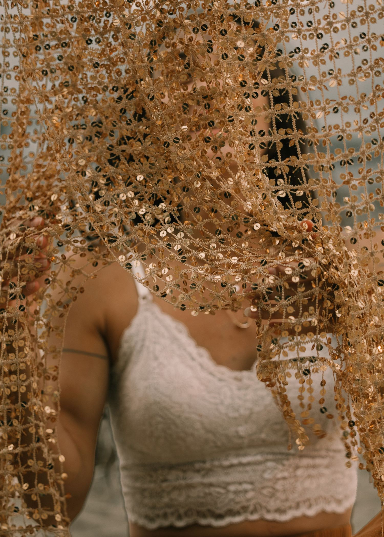 Woman in white lace top reaching through a shimmering gold sequin curtain backdrop.