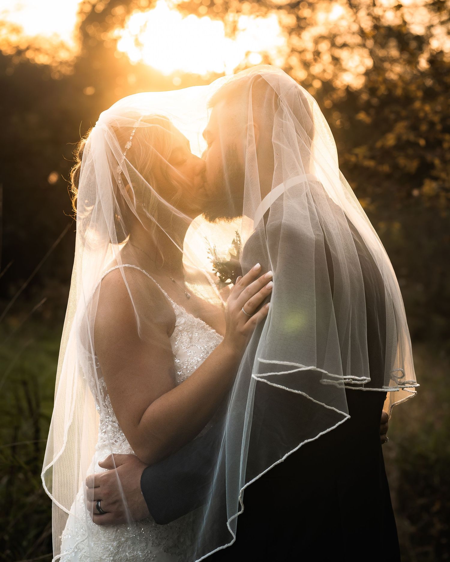 Bride and groom sharing a romantic kiss under a wedding veil at golden sunset outdoors.