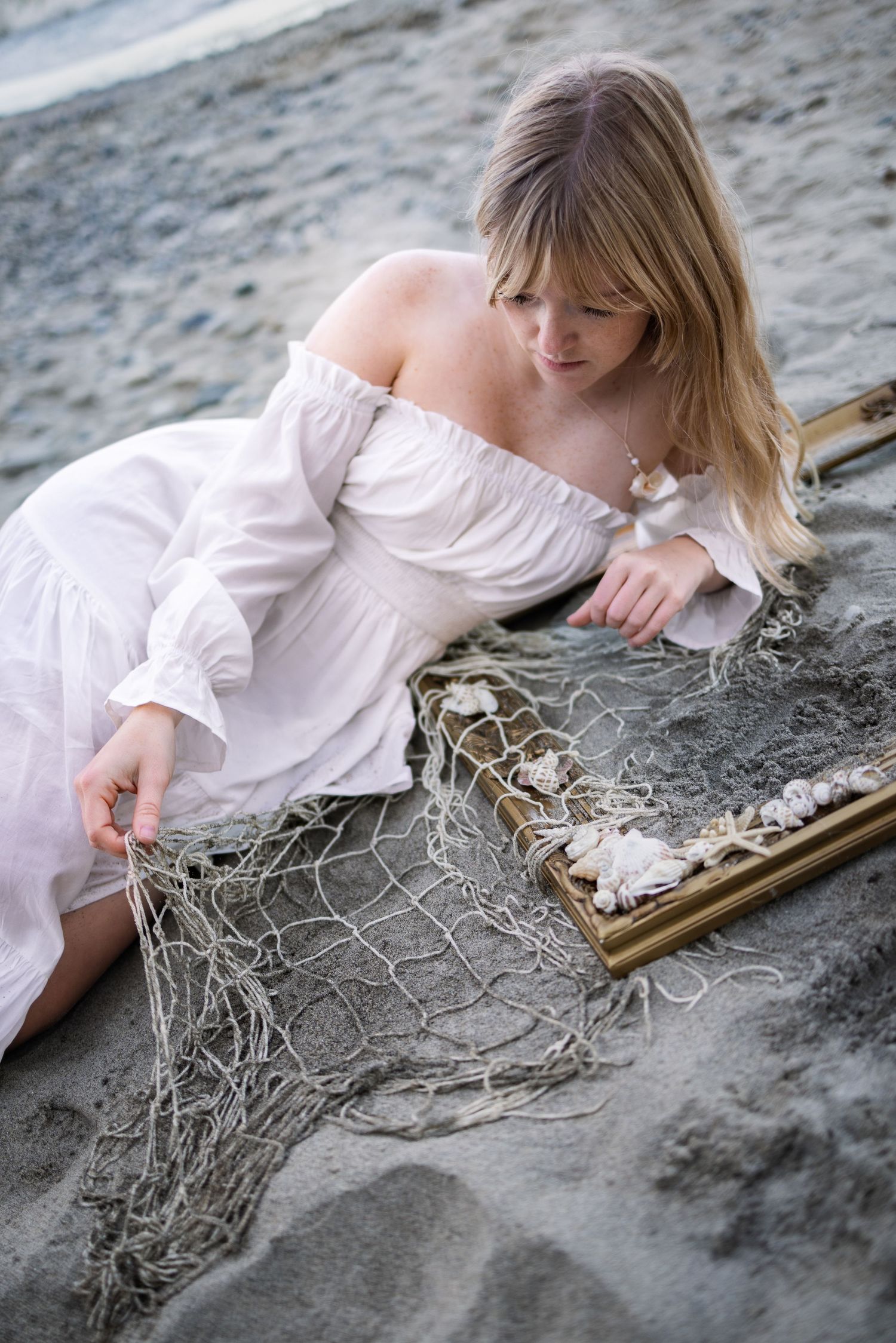 Woman in white off-shoulder dress sitting on sandy beach examining fishing net with seashells in wooden frame.