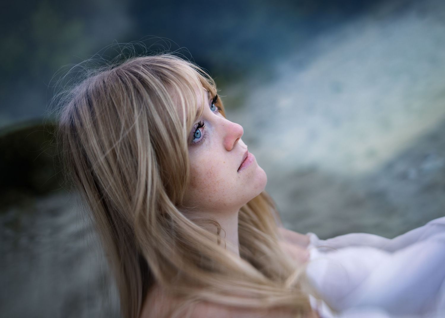 Young blonde woman with blue eyes gazing upward thoughtfully, soft bokeh background, portrait photography.