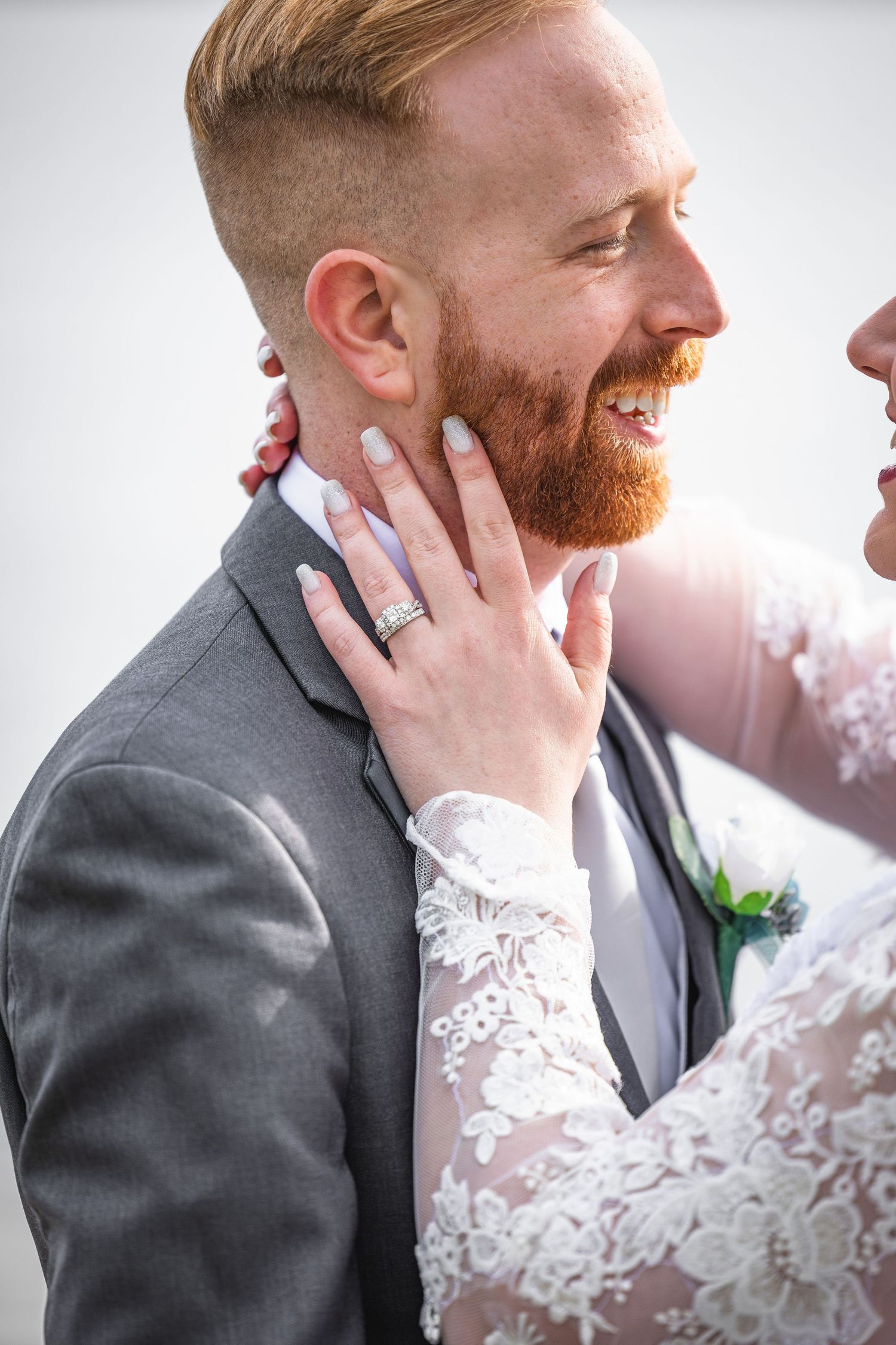 Smiling groom with red beard in gray suit as bride touches his face at wedding.