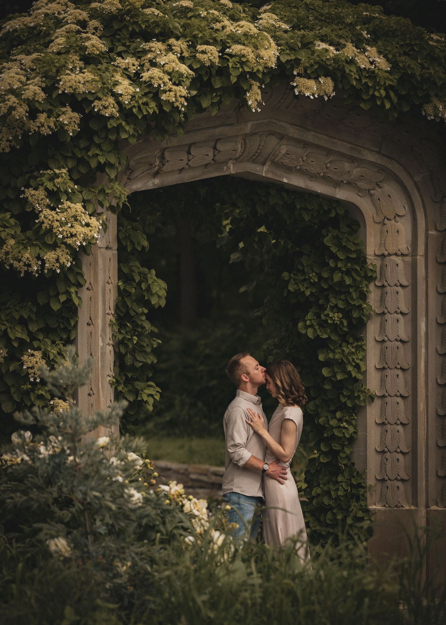 Couple sharing a tender moment under a flower-covered stone archway in a lush garden setting.