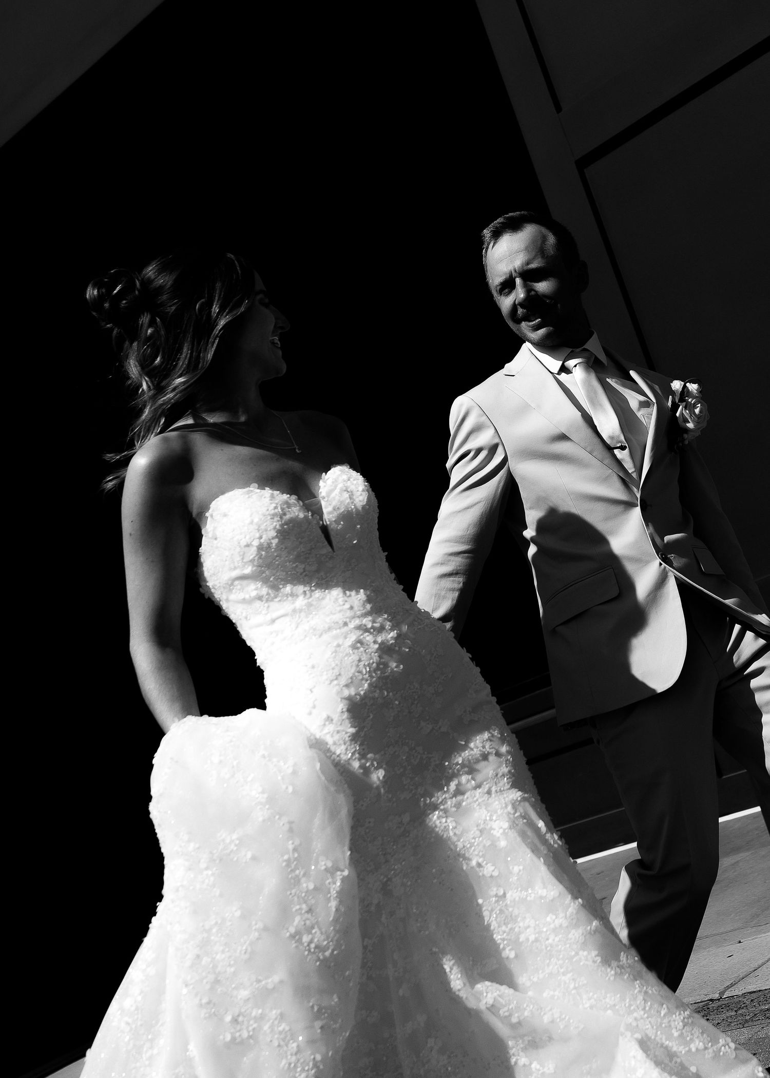 Artistic black and white shot of bride in strapless lace gown and groom in light suit walking together.
