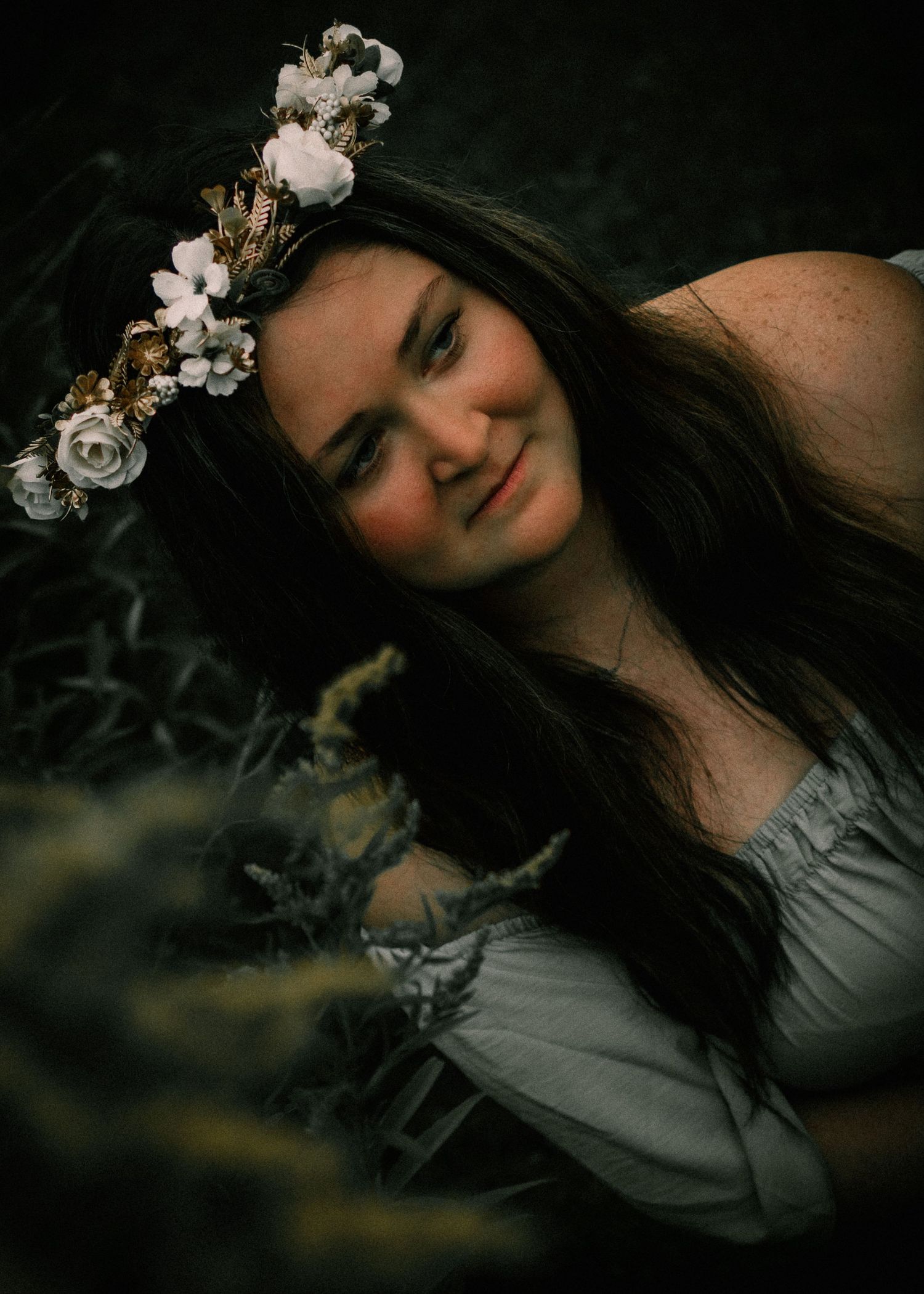 Young woman with long dark hair wearing a floral crown of white and gold roses, smiling softly outdoors.