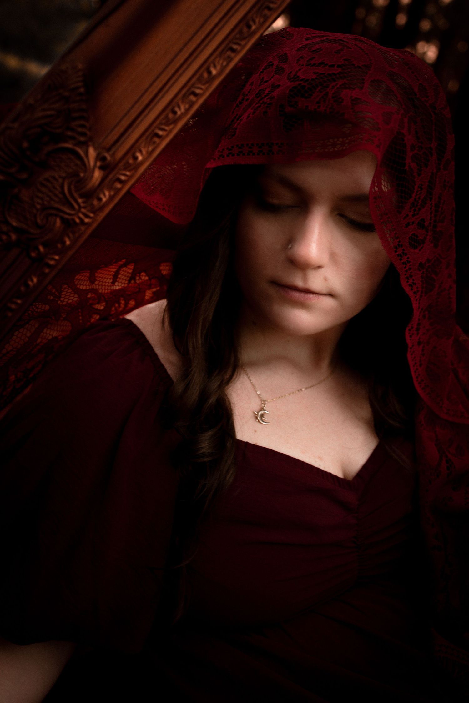 Young woman in dark red dress and lace hood looks down pensively, wearing a crescent moon necklace.