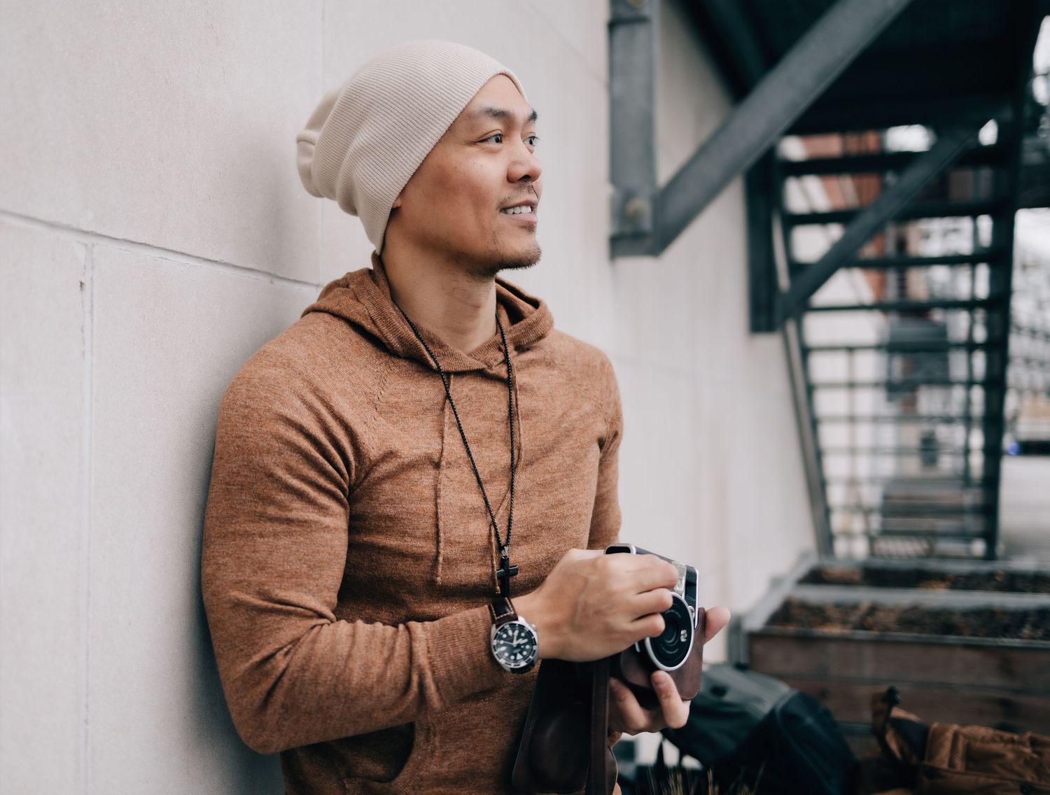 Stylish Asian man in beanie and brown hoodie leaning against wall holding coffee outdoors.