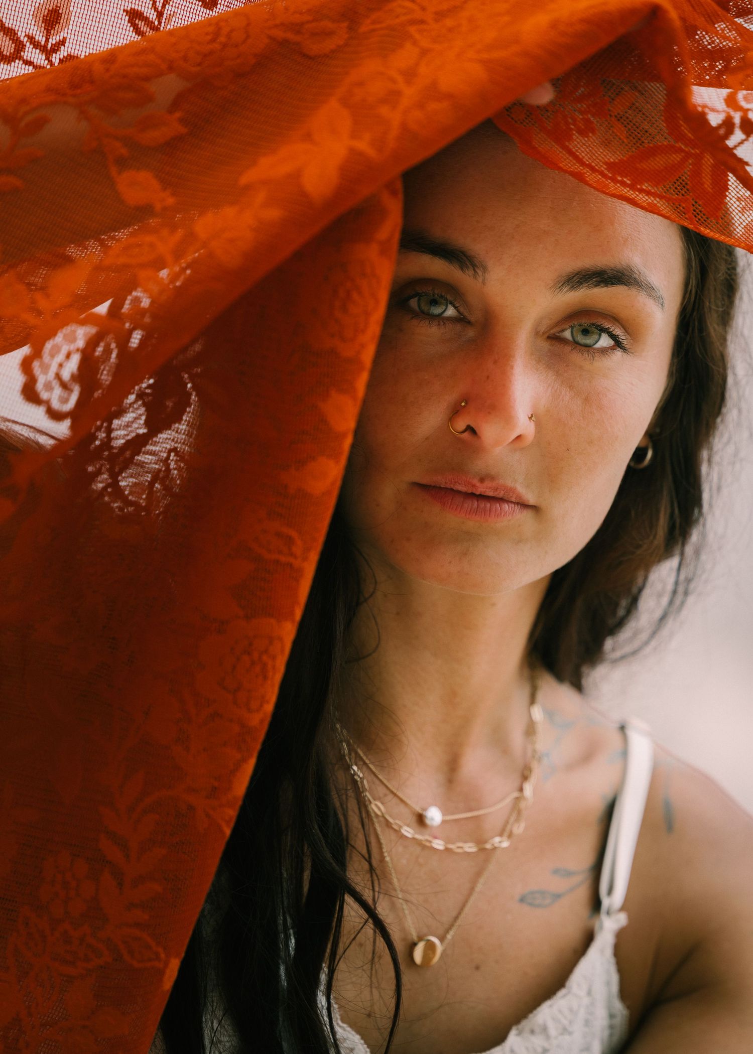 Woman with green eyes partially covered by vibrant orange lace fabric, wearing layered gold necklaces.
