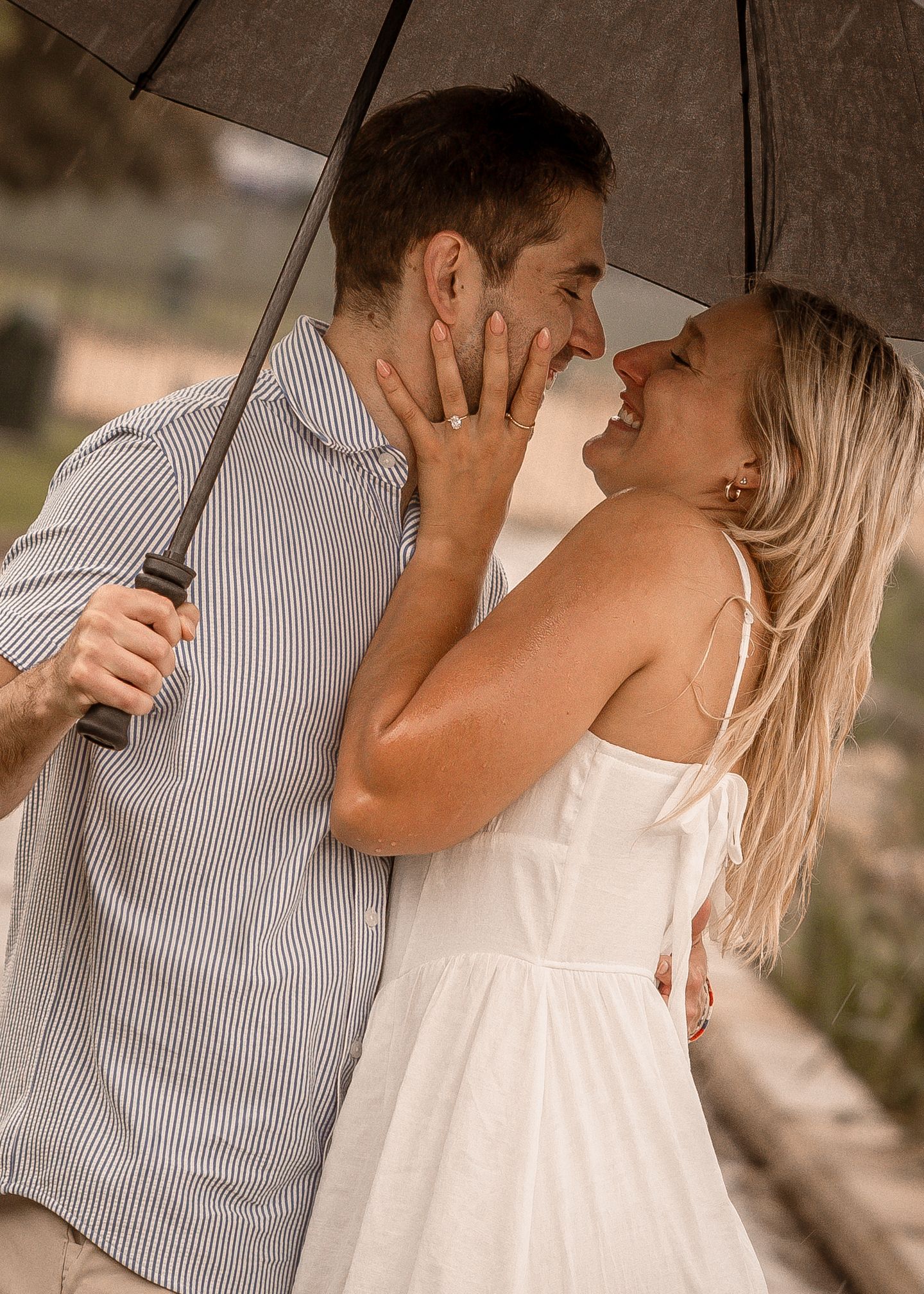 Couple sharing a romantic moment under an umbrella, woman in white dress smiling as they nearly kiss.