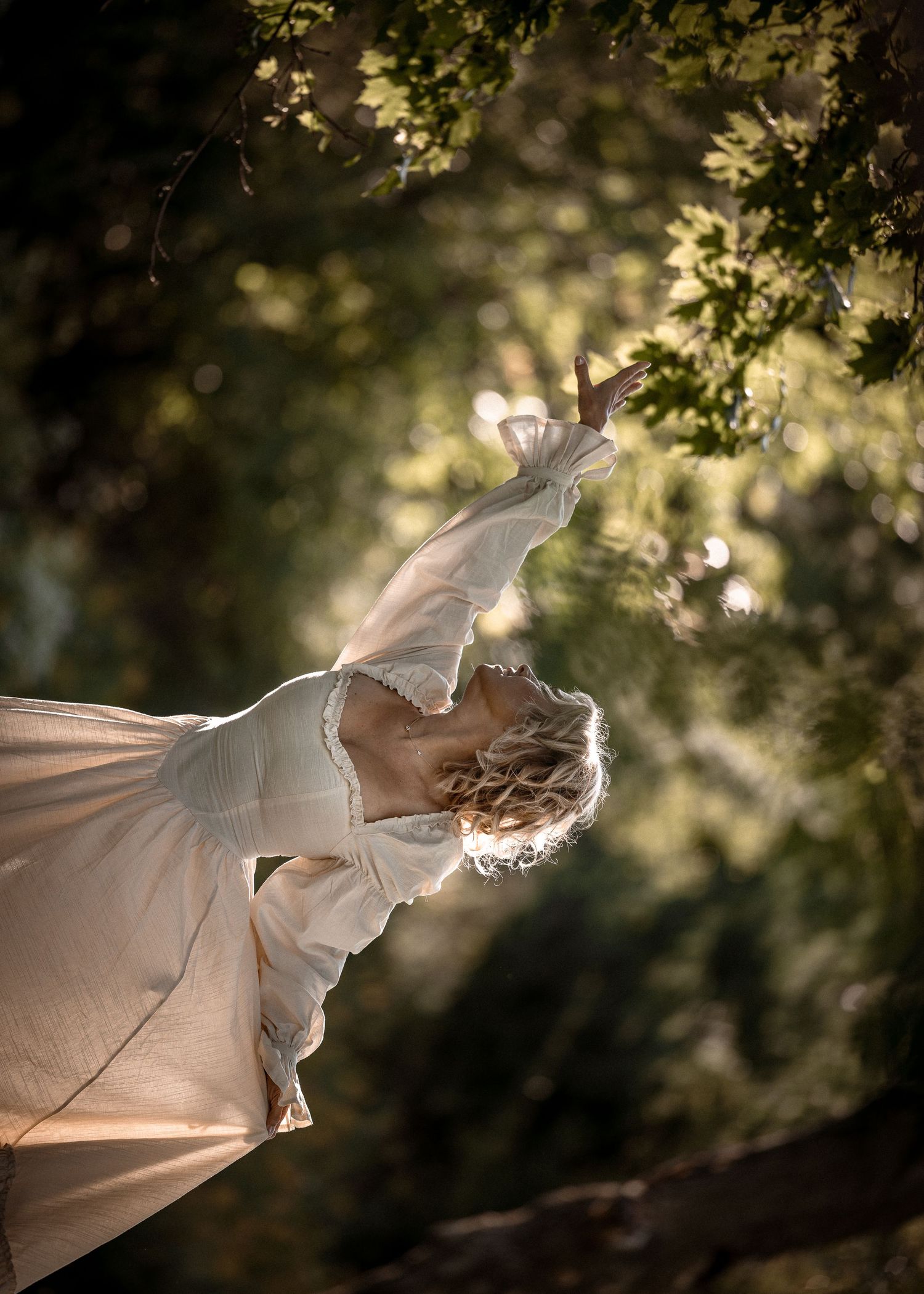 Woman in white dress reaching toward sunlit tree branches in a dreamy forest setting.