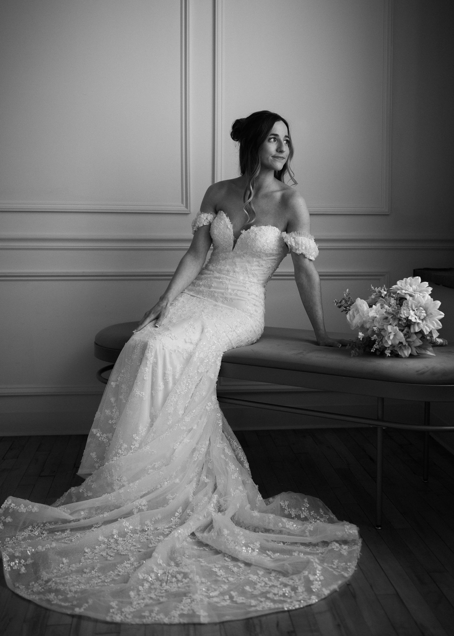 Elegant bride in off-shoulder lace mermaid gown seated with bouquet in a classic black and white portrait.